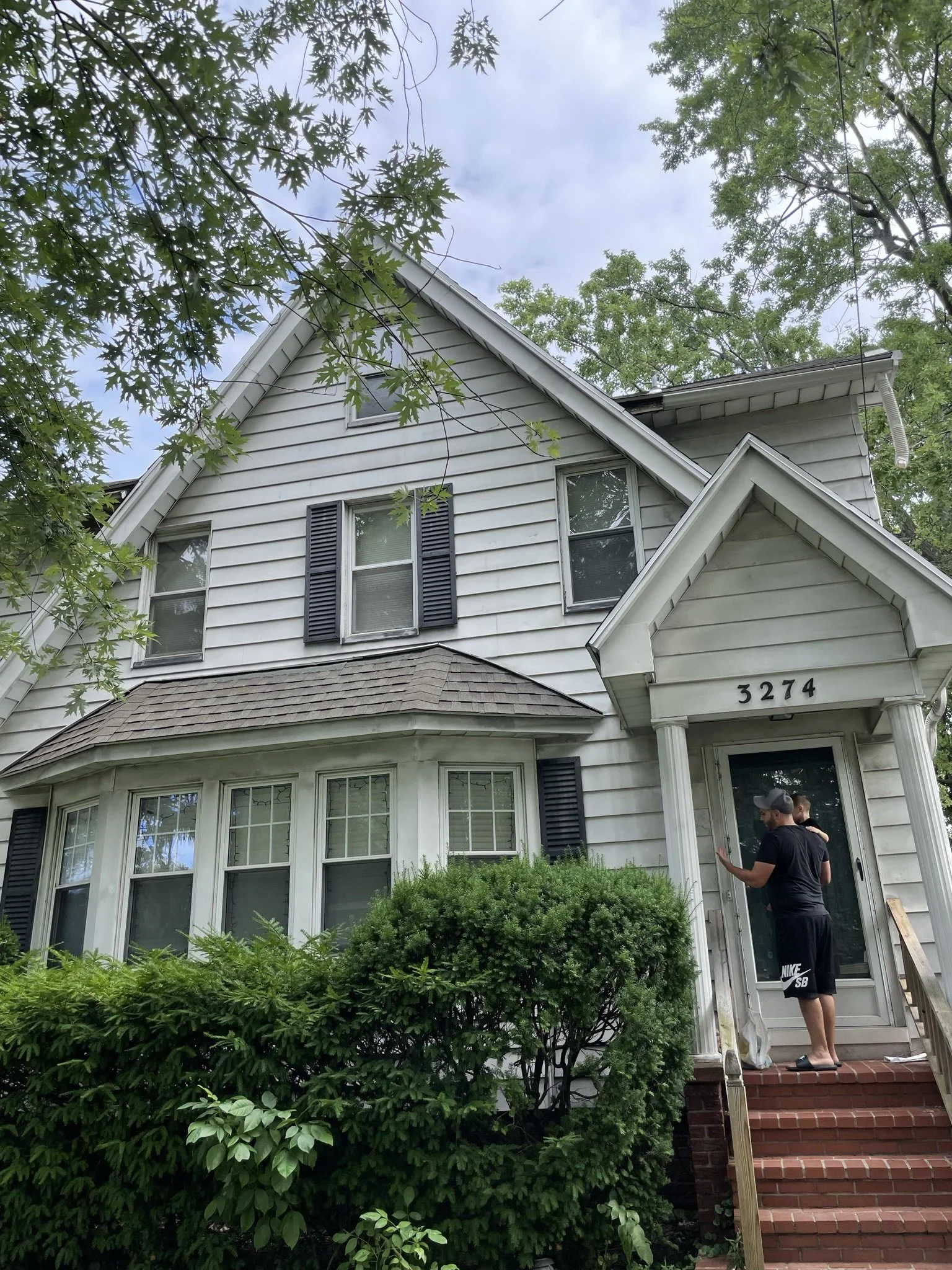 A white, three-story house with black shutters, a front porch, and steps leading to the entrance. Two people are standing on the porch near the door, one person is wearing a black shirt and shorts, and the other is partially visible. The house is surrounded by green bushes and trees, with a cloudy sky overhead.
