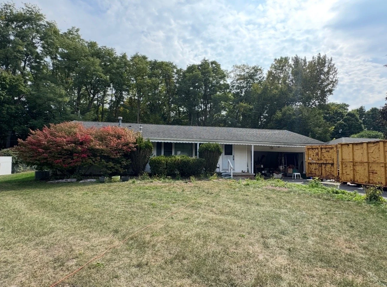 A single-story house with a gray roof and white siding, surrounded by grass and trees, with a large shrub with pink flowers in the front yard and a wooden fence on the right side.