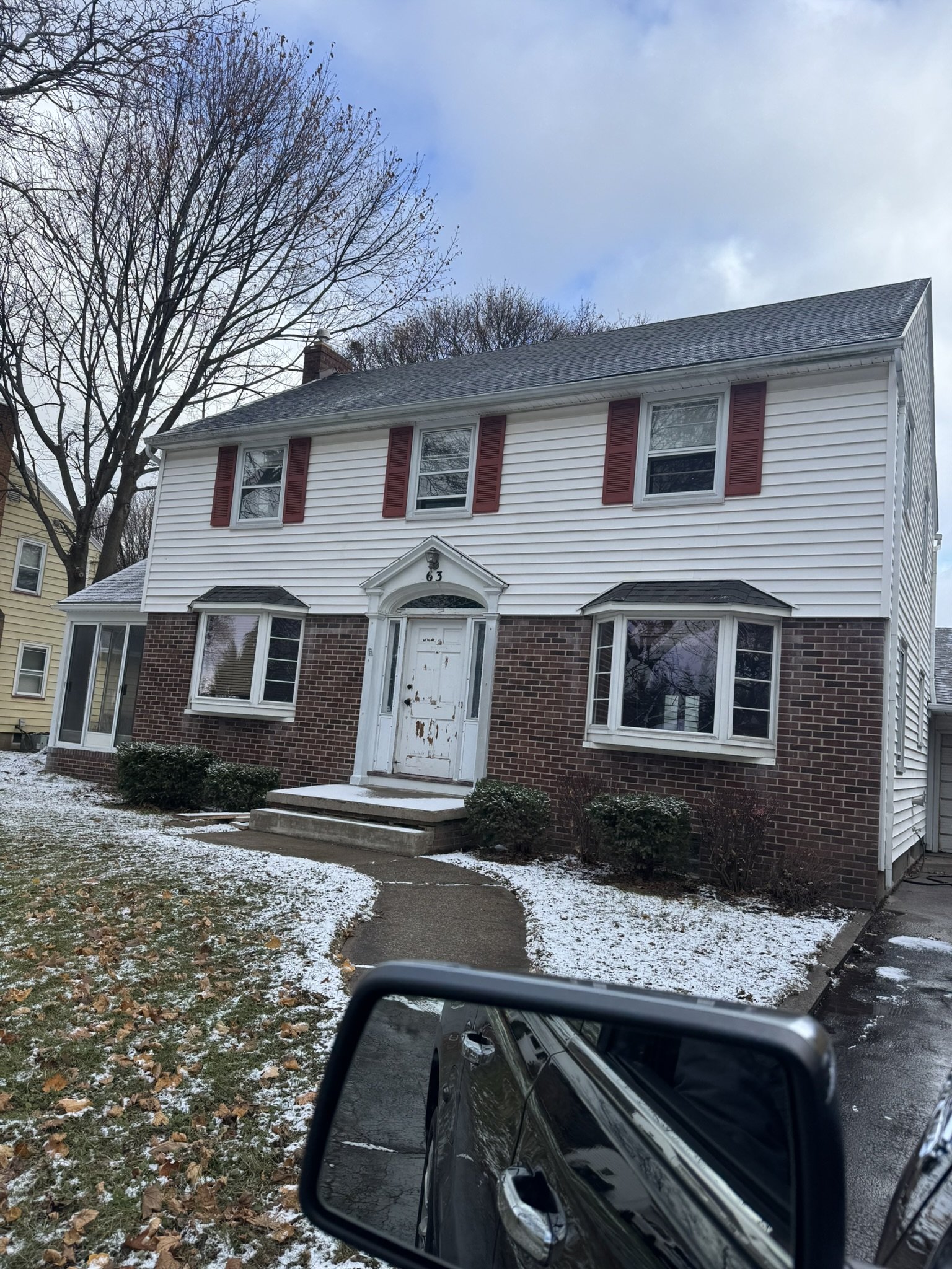 A two-story house with white siding on the upper level and red brick on the lower level. The house has five windows, some with black awnings and red shutters, and a front porch with steps leading to a white front door.