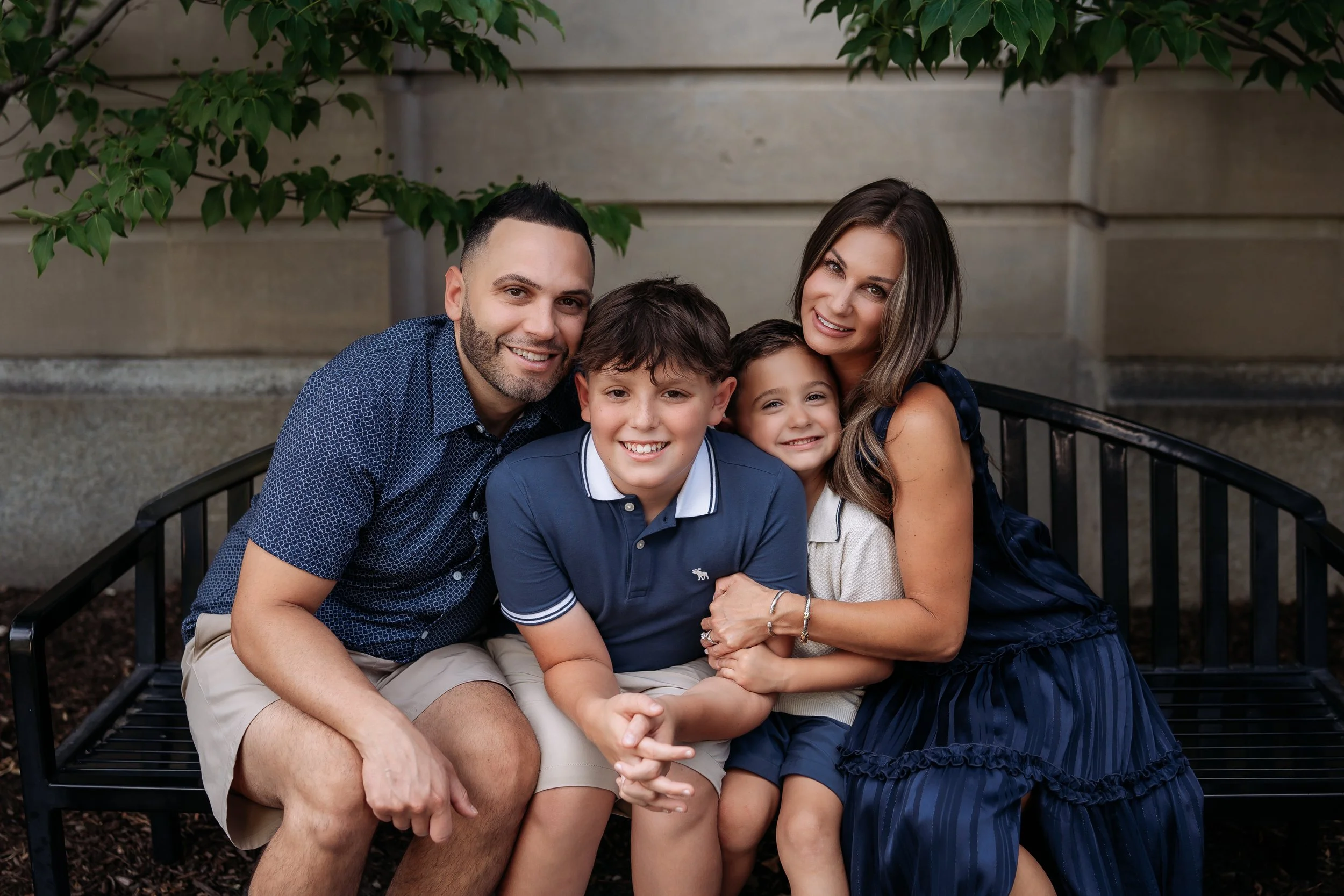 A family of four sitting on a black metal bench outdoors, smiling at the camera. The family includes a man, woman, and two children, a boy and a girl.