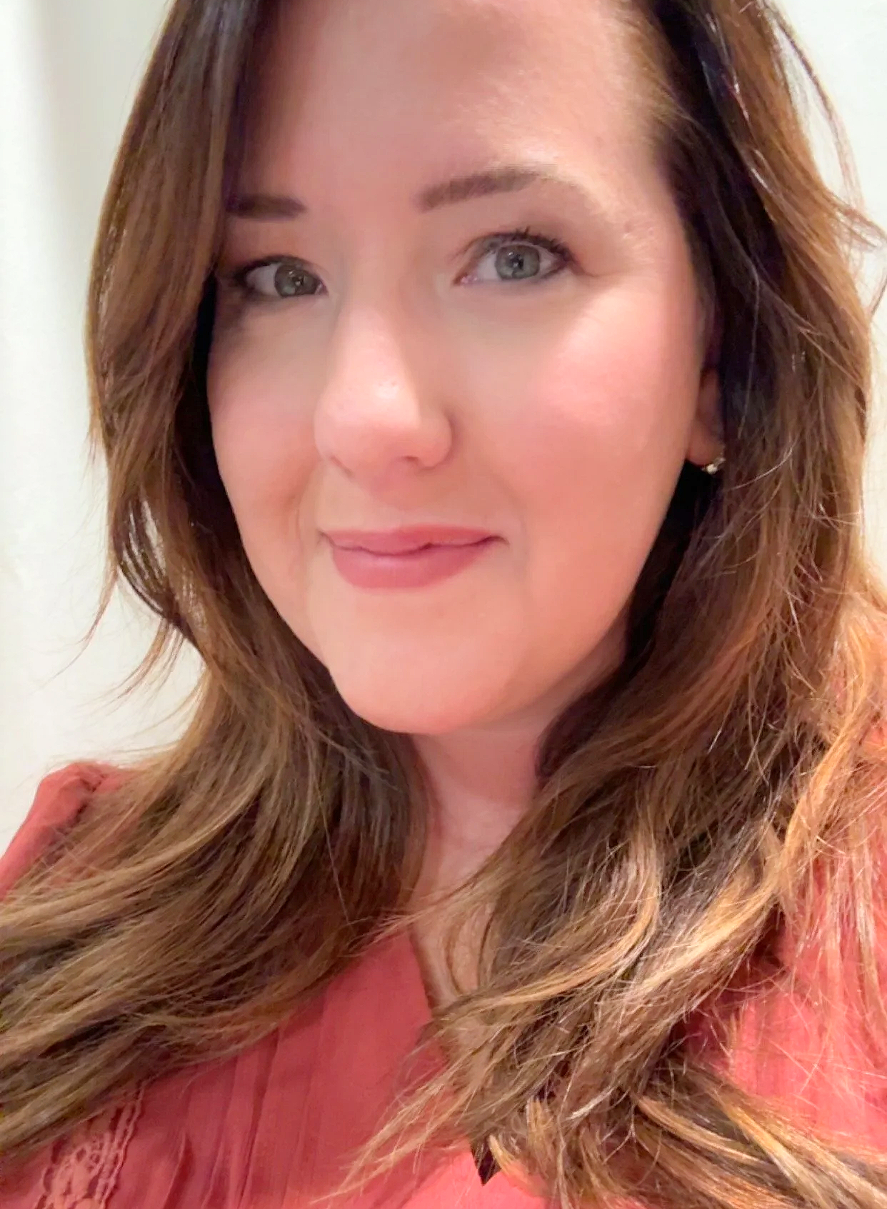 Close-up of a woman with long, wavy brown hair, light makeup, and a pink top, smiling softly.