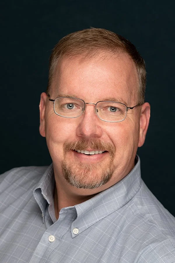 A smiling middle-aged man with glasses, short light brown hair, and a beard wearing a light gray button-up shirt against a dark background.