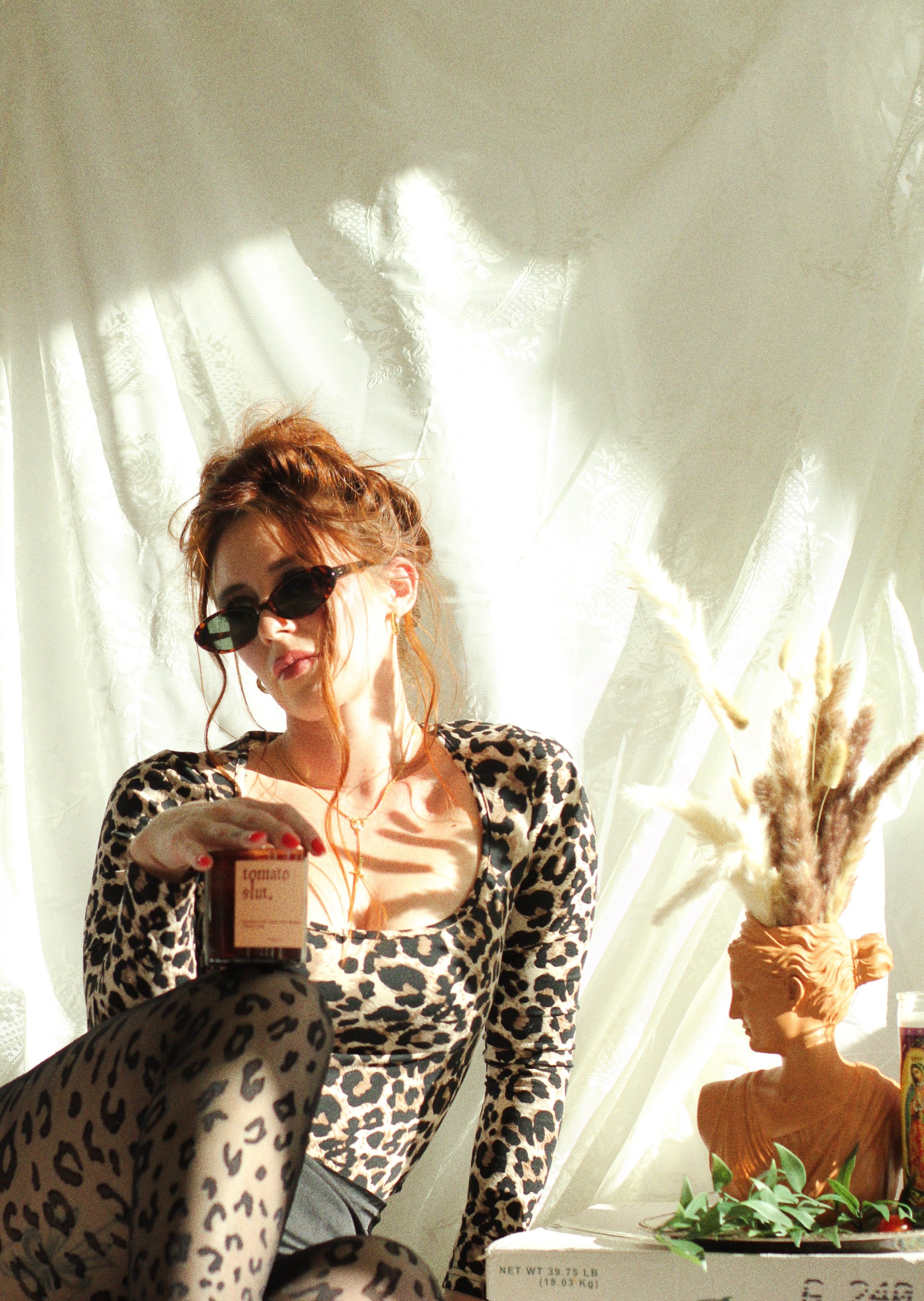 A woman with red hair, wearing leopard print clothing and sunglasses, sitting in front of a white curtain with sunlight shining through. She is holding a small carton of juice. There is a sculpture of a person's head with plant-like feathers on top, 