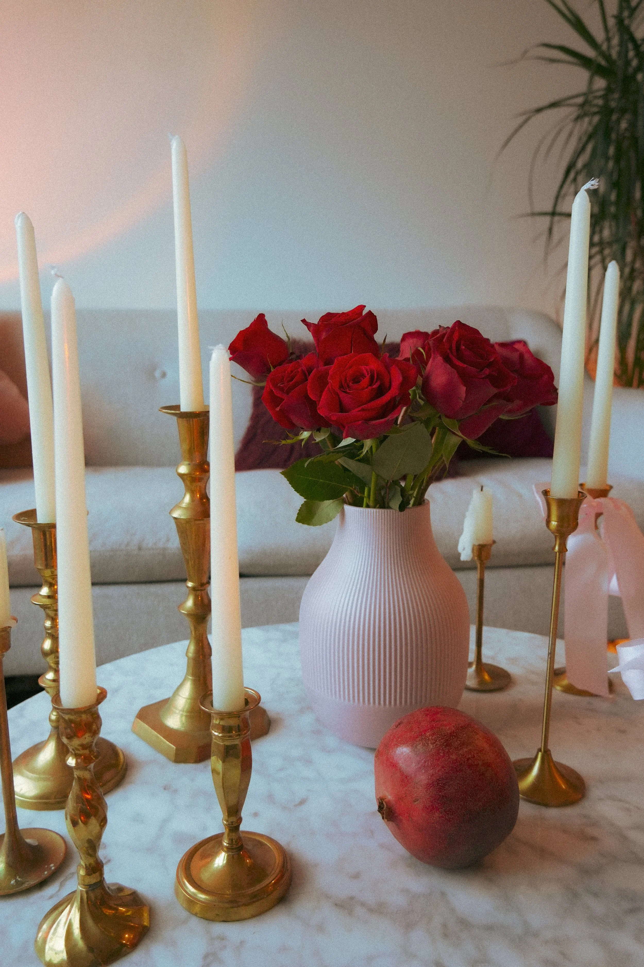 A table with a pink vase of red roses and several golden and white candles in candleholders, next to a pomegranate, in a cozy living room.