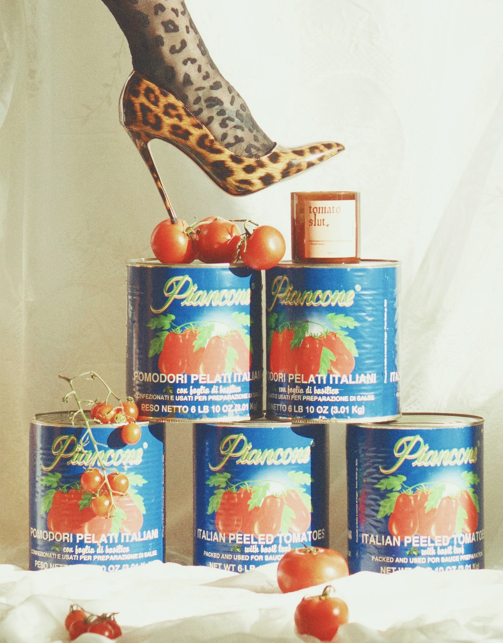 Stack of canned peeled Italian tomatoes with a bunch of fresh tomatoes on top, a small cherry tomato on a wire, a single tomato at the front, a high-heeled leopard print shoe stepping on the cans, and a small jar labeled 'tomato slit' in the background.