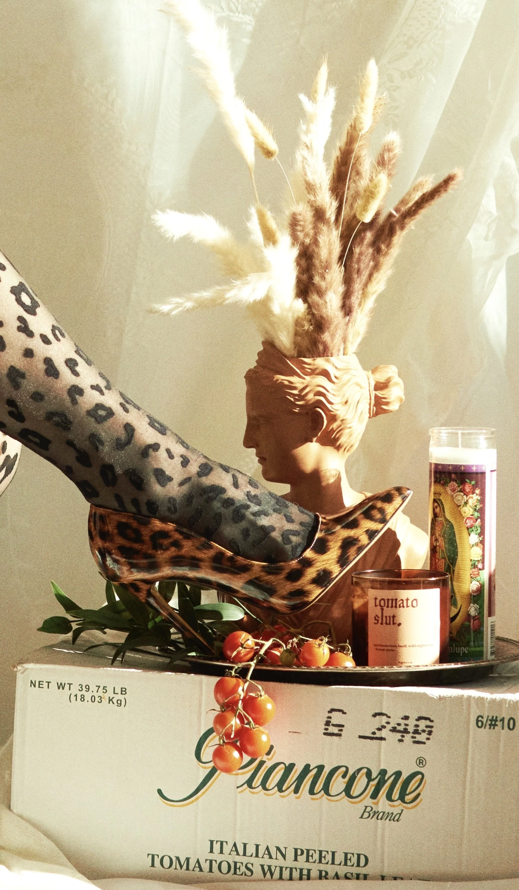 Decorative arrangement featuring a bust with a hairdo and pampas grass, leopard print shoes, a leopard print stocking, a tomatoes and greenery, a candle, and a religious figure on a box labeled 'Giancone' brand, with a lace curtain in the background.