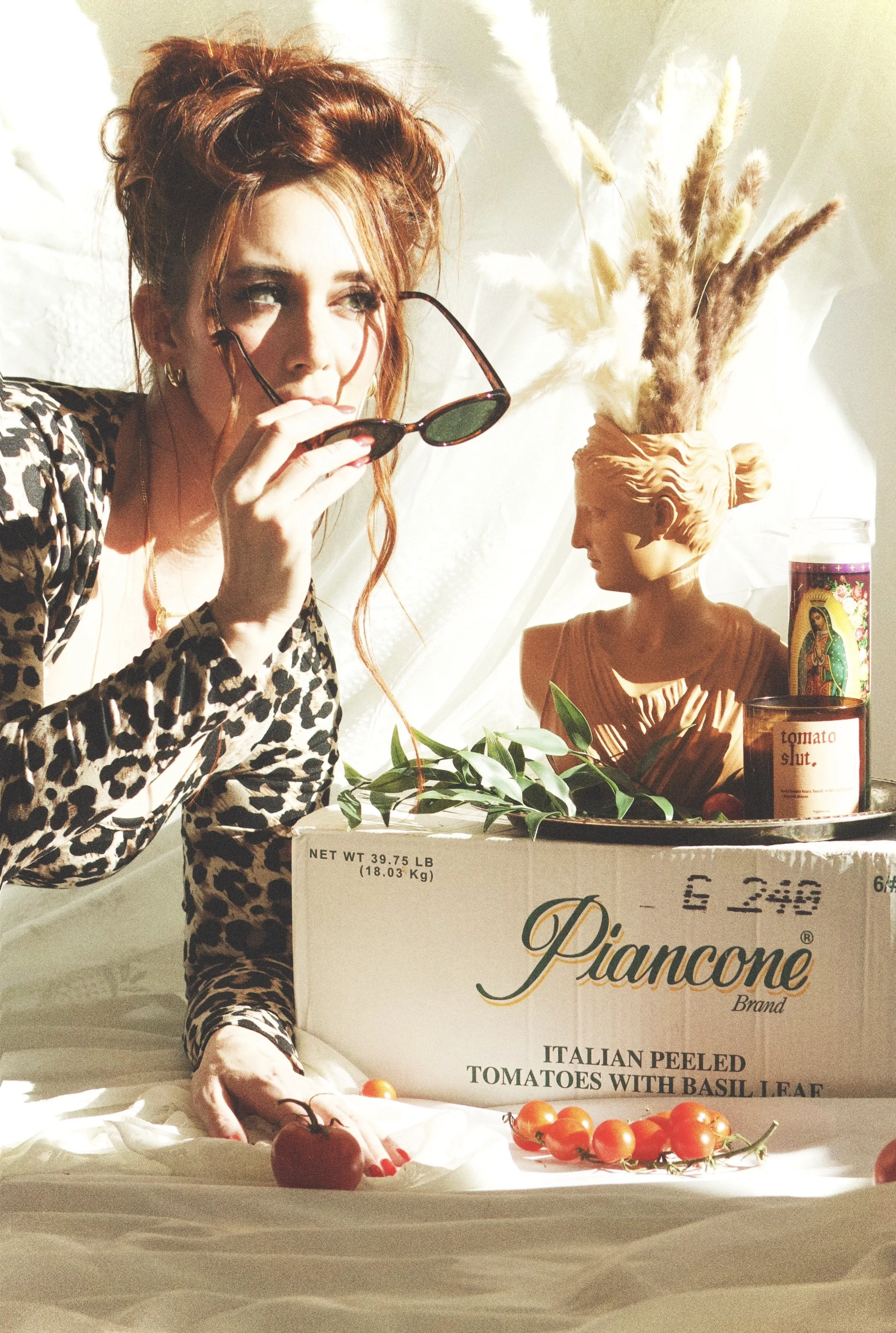 A woman with red hair and animal print clothing holding sunglasses, sitting next to a box of Italian peeled tomatoes with basil leaves, a small bust, a candle, and a container of religious candles, all illuminated by sunlight.