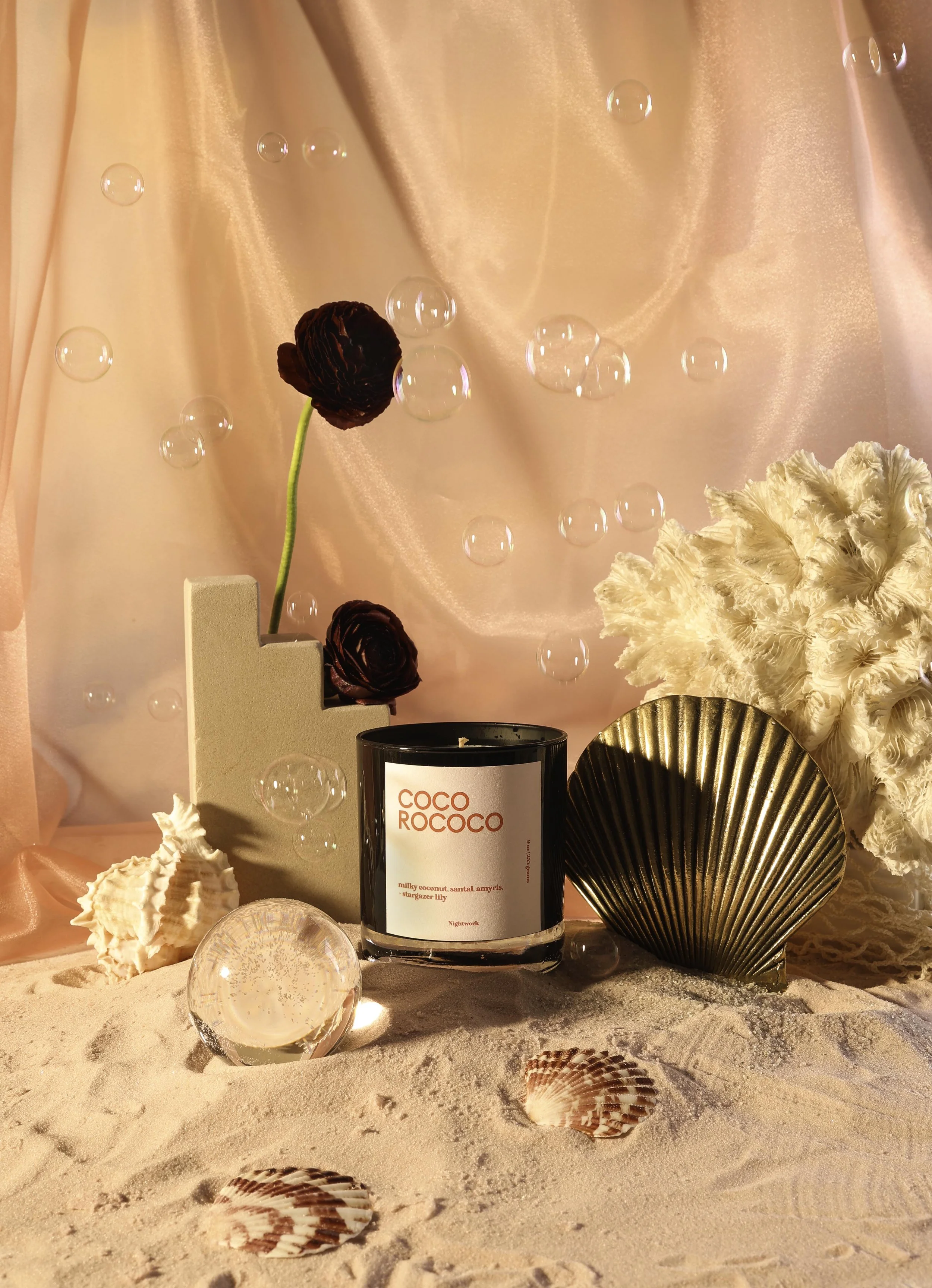 A decorative still life with a candle labeled 'Coco Rocco', seashells, a white coral, a gold fan, dark red flowers, and a clear glass sphere on a sandy surface, with soft pink fabric background and floating bubbles.