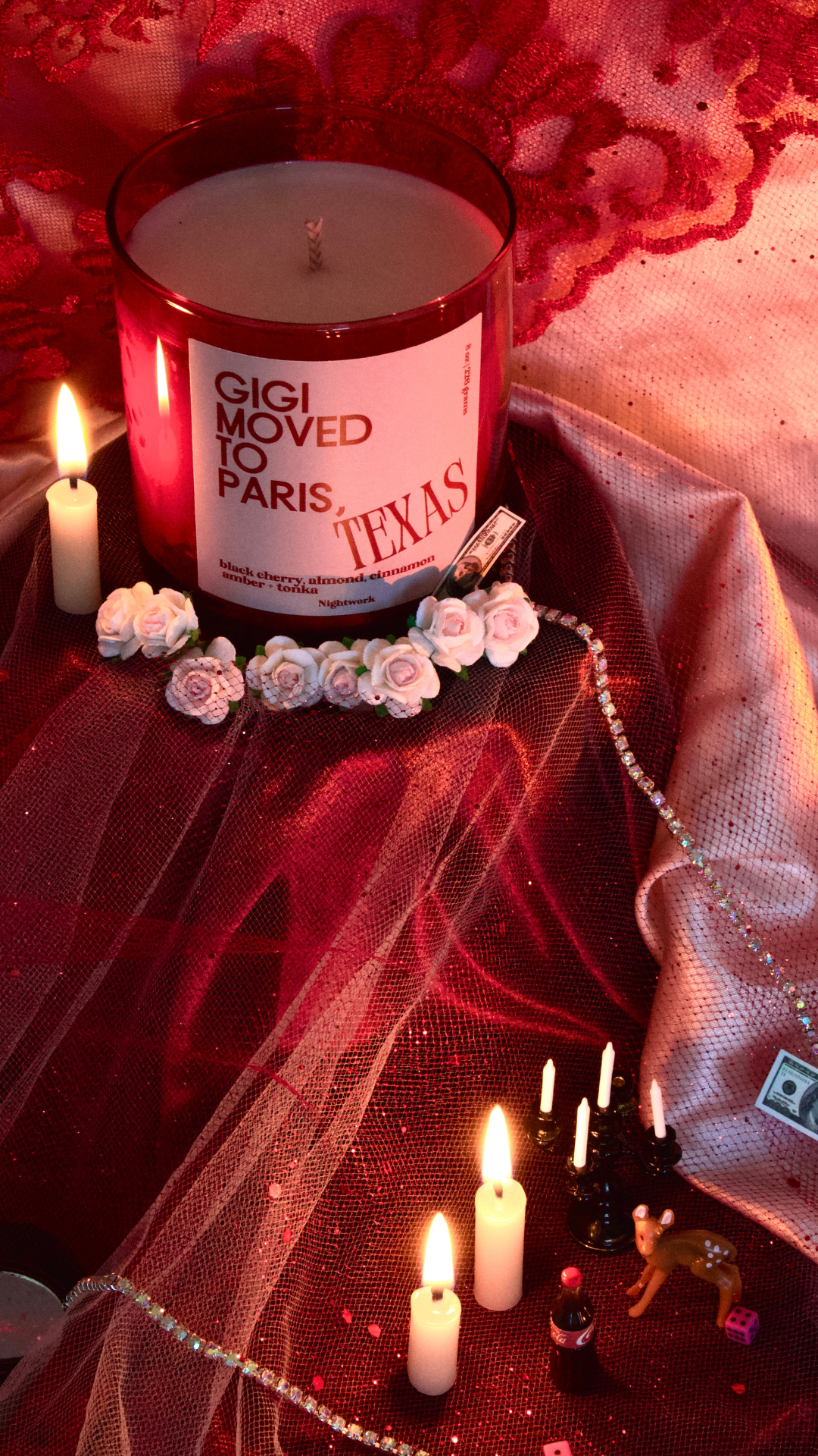 A candle in a red glass jar labeled 'Gigi moved to Paris, Texas' surrounded by fake flowers, small candles, a deer figurine, tiny bottles, and pink dice on red and pink fabric.