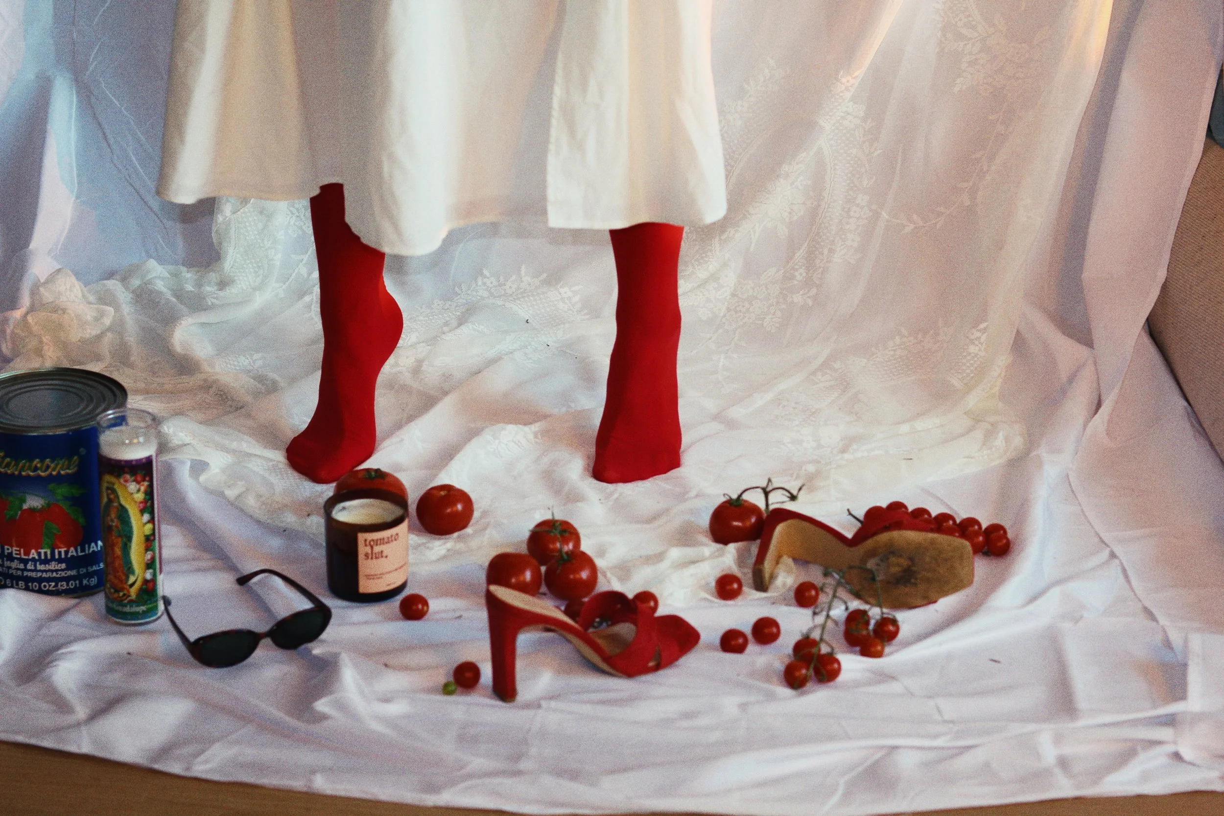 Red high-heeled shoe, fresh tomatoes, sunglasses, canned food, candle, and a shoe on a white fabric backdrop with a white dress and curtain.