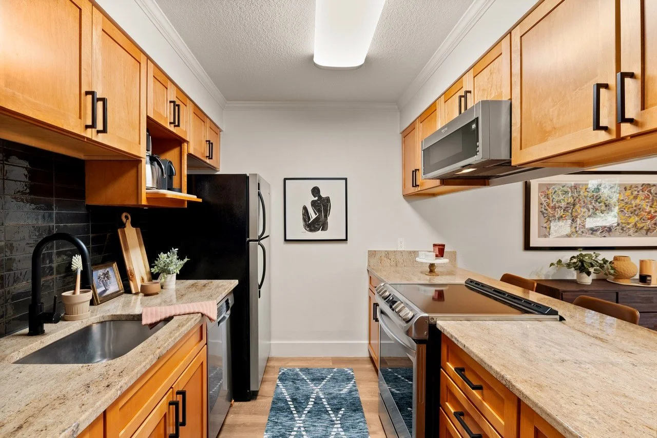 Kitchen with wooden cabinets, granite countertops, black backsplash, stainless steel appliances, black fridge, black microwave, and black faucet. Decor includes framed artwork, plants, and small decorative items.