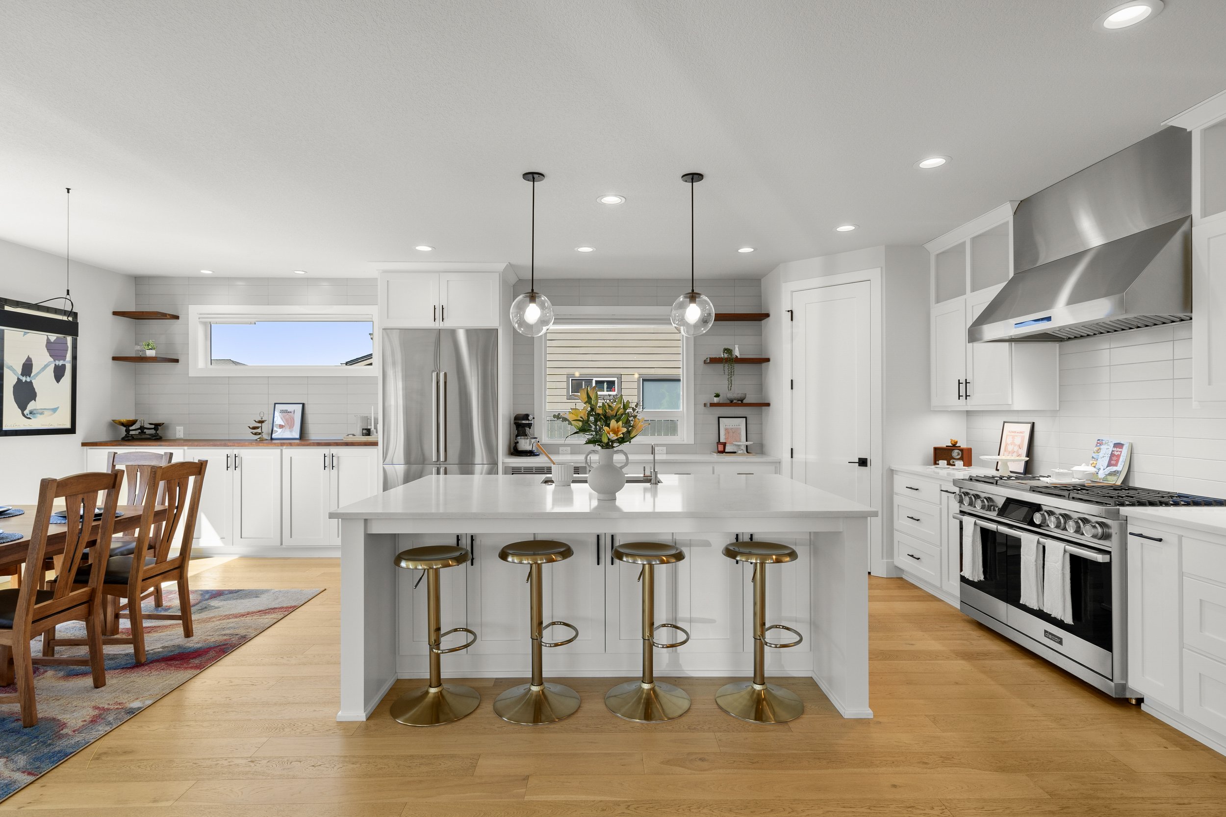Modern white kitchen with a large island, four gold bar stools, stainless steel appliances, open shelving, and decorative accents.