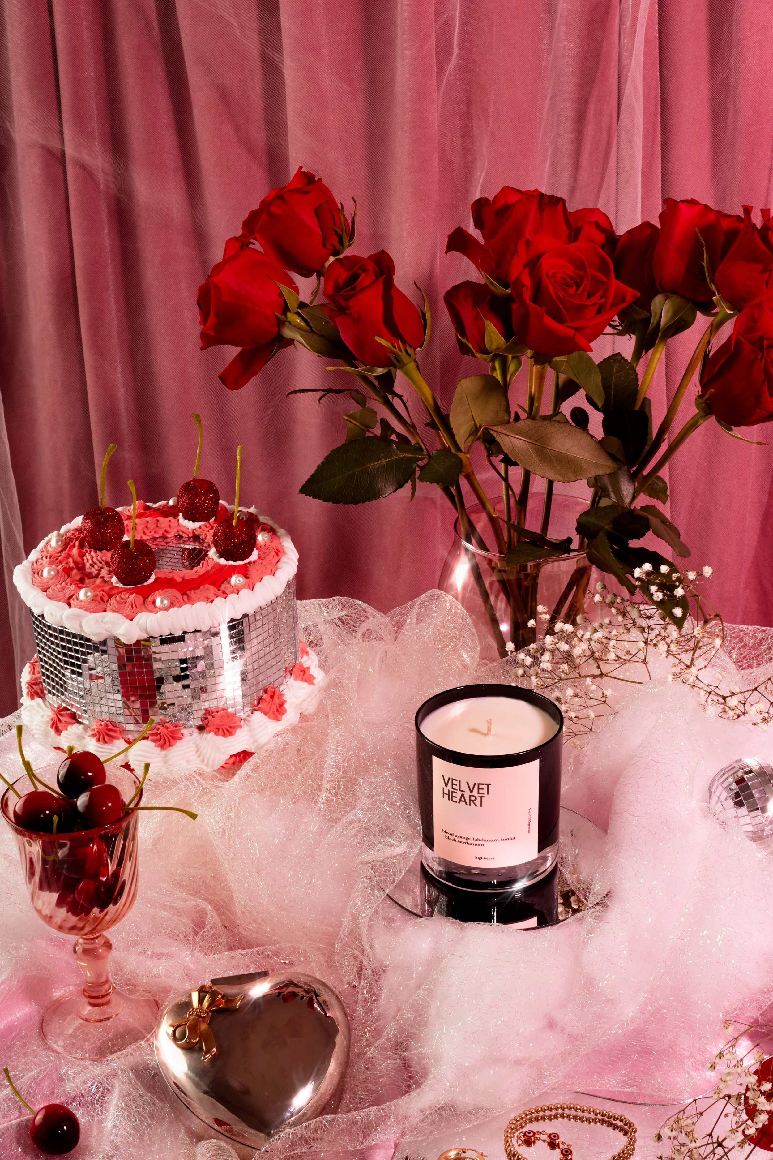 A pink background with a table decorated with a bouquet of red roses, a heart-shaped mirror, a glass of cherries, a pink cake with cherries and pearls, a white candle labeled 'Velvet Heart', and gold jewelry, all surrounded by pink decorative gauze.