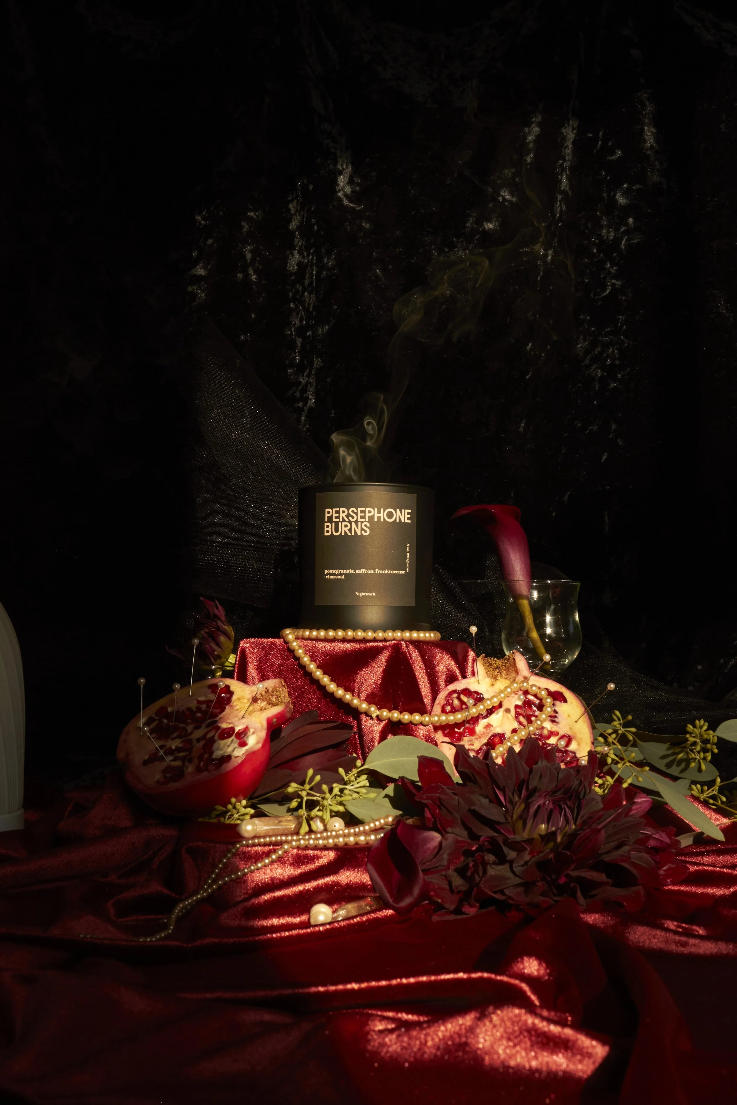 A dark, artistic still life with a black candle labeled "Persephone Burns," pomegranate halves, a clear glass, pearl necklaces, dark red flowers, and greenery on a red velvet fabric.