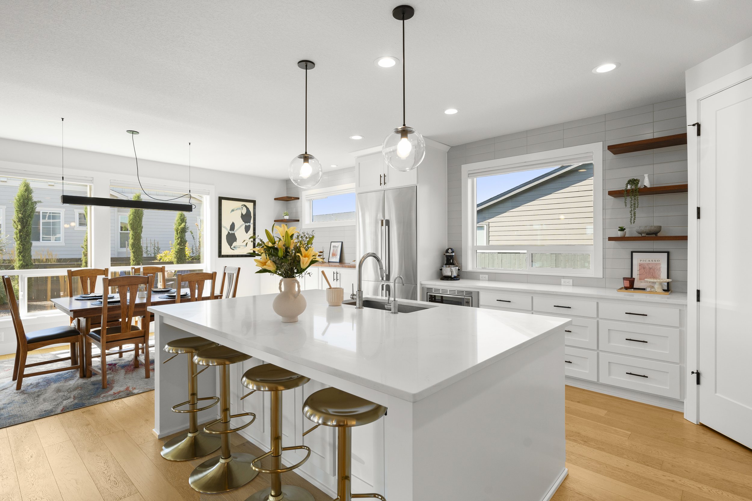 Modern kitchen with white cabinets and island, wooden stools, flowers in a vase, dining area with wooden table and chairs, large windows with views, floating shelves, and hardwood floors.