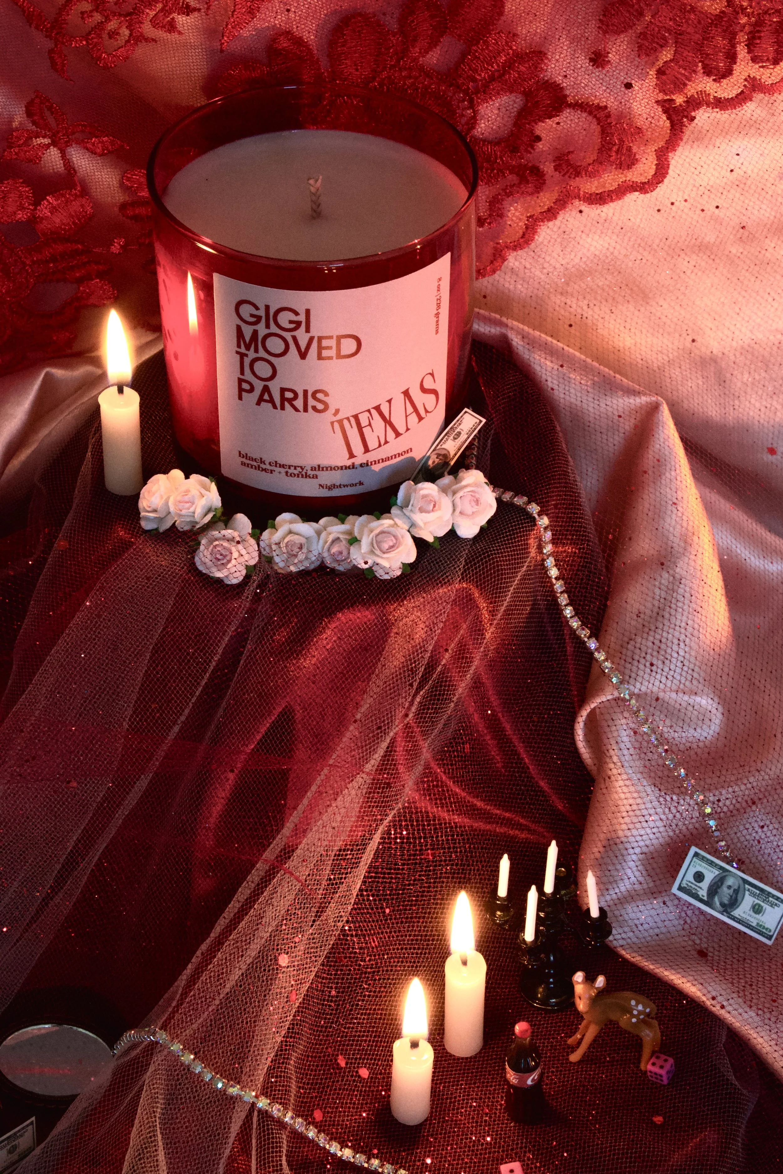 Decorative setup featuring a large red candle with a label that reads 'Gigi moved to Paris, Texas,' surrounded by miniature candles, artificial pink roses, a small deer figurine, a tiny bottle, and a fake dollar bill, all arranged on red and pink fab