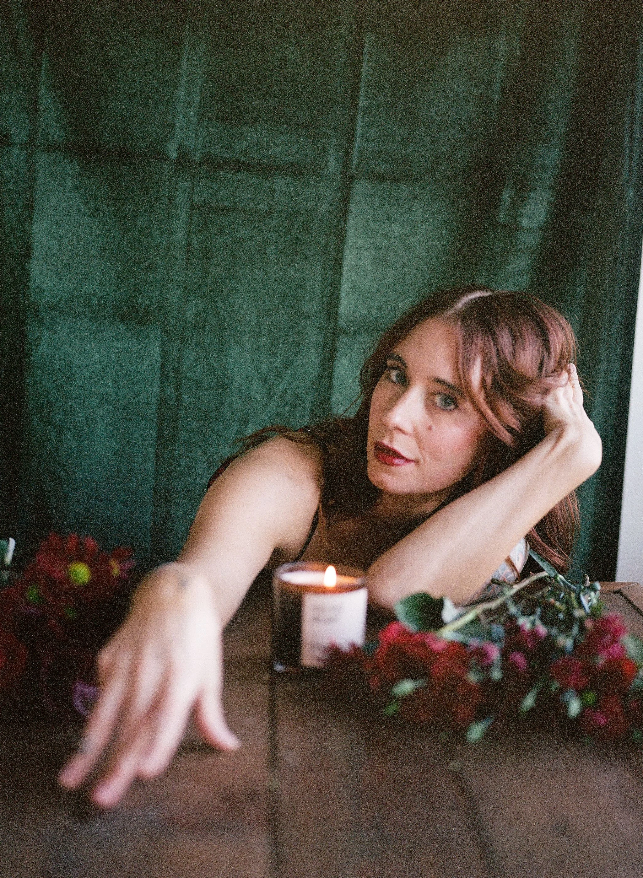 A woman with red hair and makeup poses with her arm extended on a wooden table, surrounded by a lit candle and red flowers, against a dark green background.