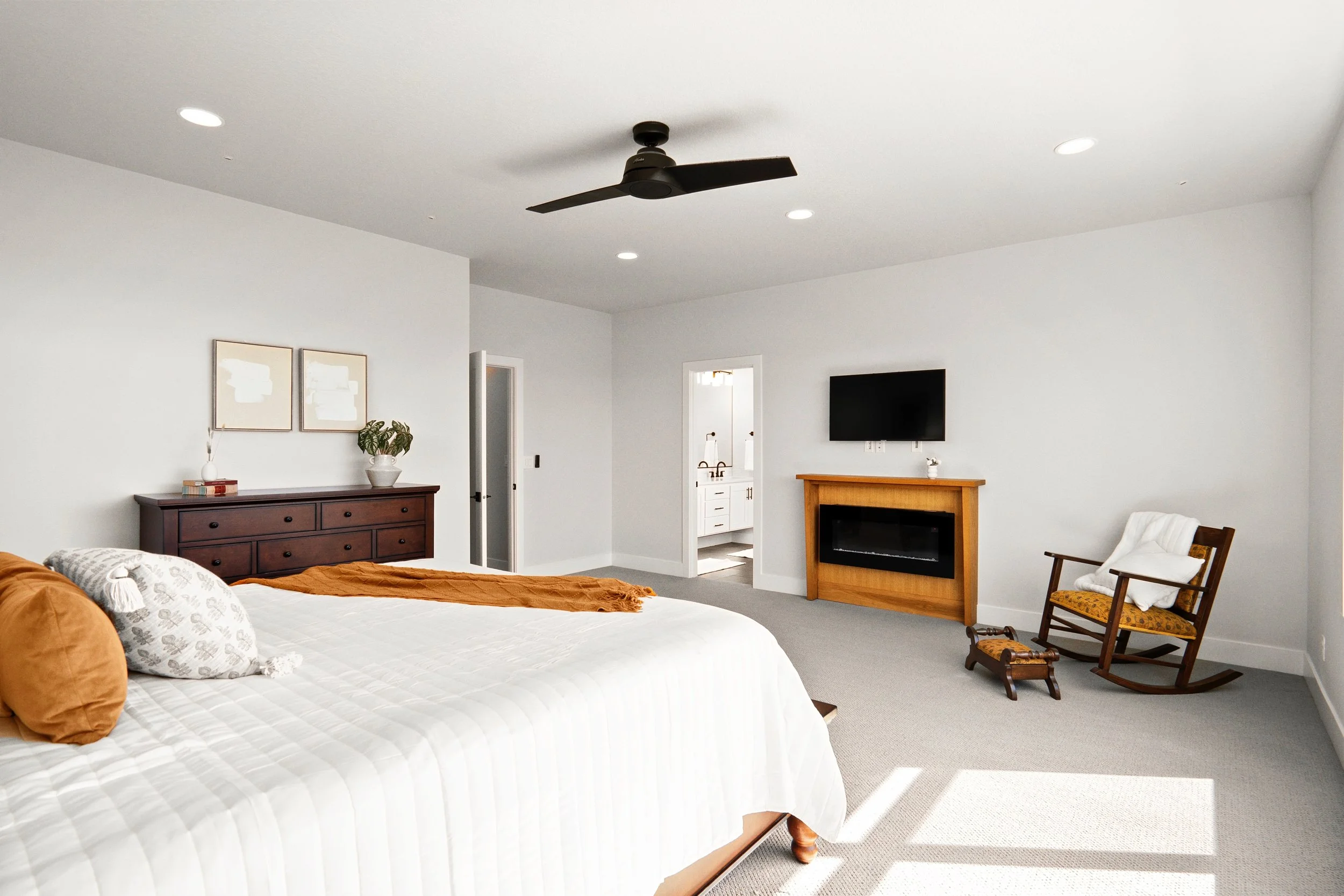 Bright and spacious bedroom with a large bed, wooden furniture, a wall-mounted TV above a fireplace, a rocking chair with a blanket, and a view into an attached bathroom.