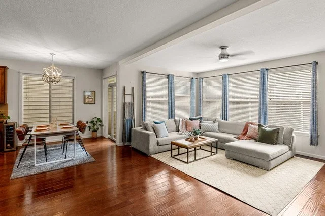Living room with large sectional sofa, coffee table, and windows with blue curtains; adjacent dining area with table and chairs, hardwood floors, and modern light fixture.