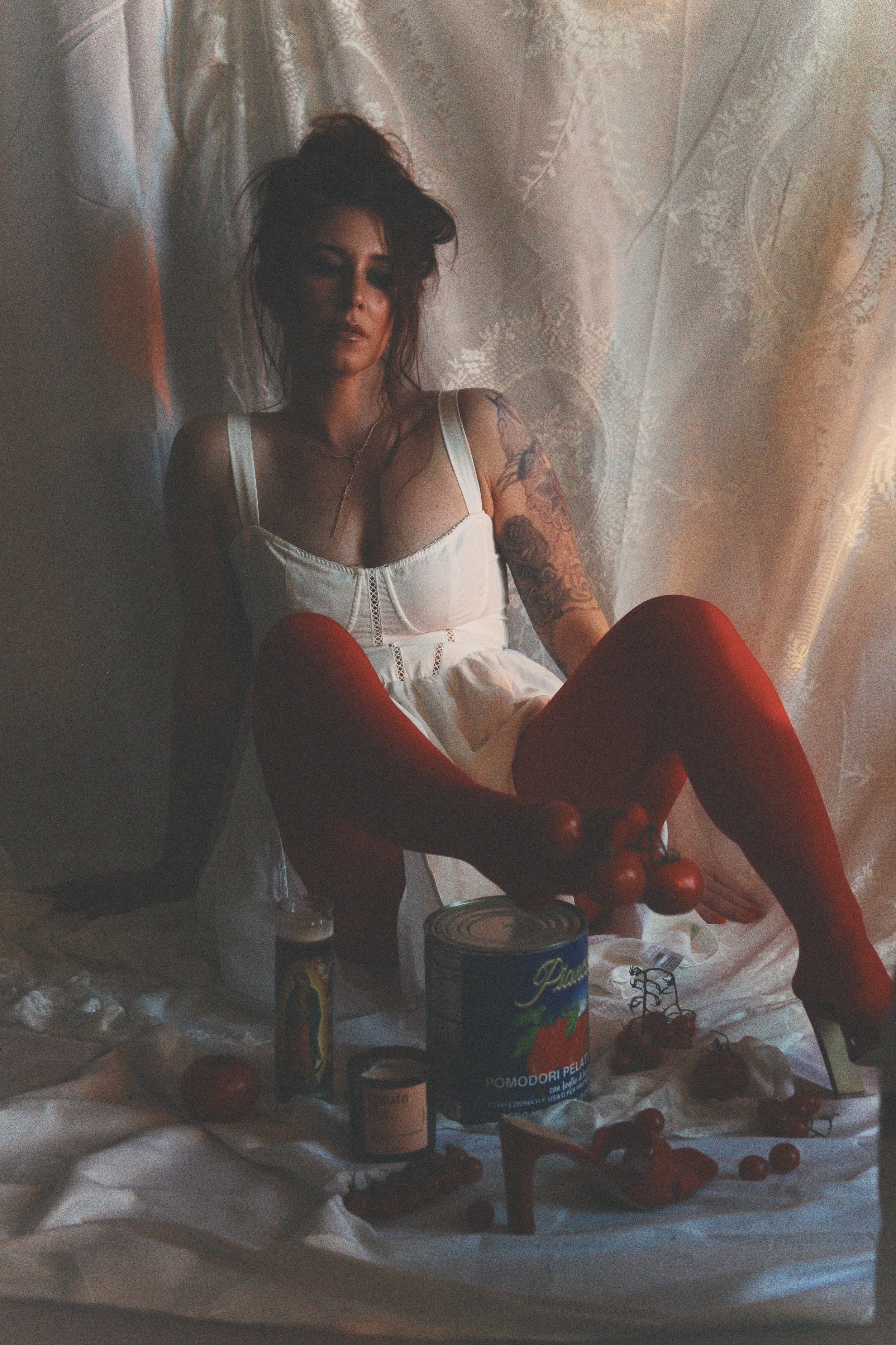 A woman with tattoos wearing a white dress and red stockings, sitting on the floor surrounded by canned tomatoes, a bottle, tomatoes, and red high-heeled shoes, in front of lace curtains.