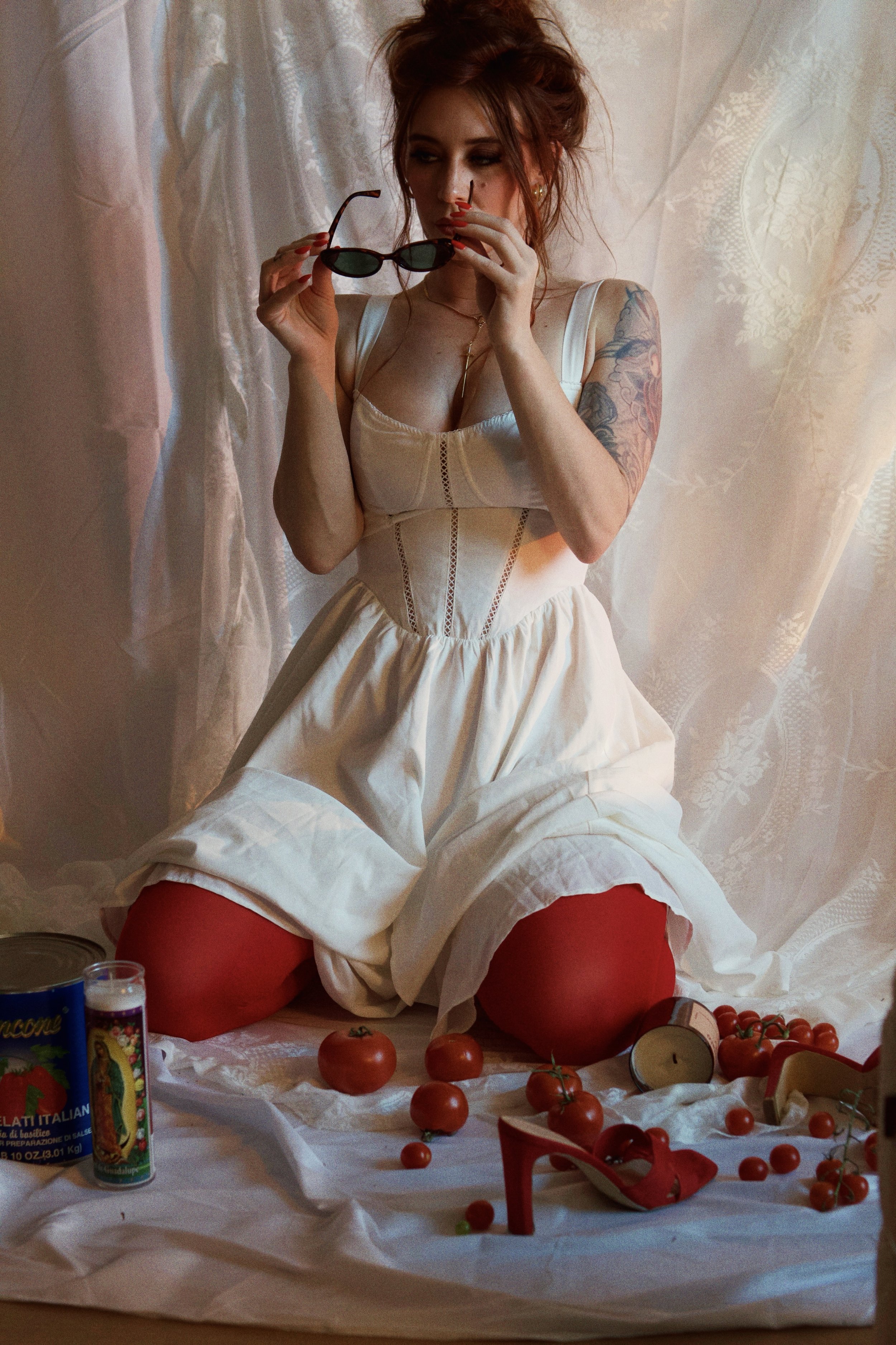 A woman with red hair holding sunglasses, seated on a white sheet surrounded by tomatoes, a candle, and a red high heel shoe, with a light-colored lace curtain background.