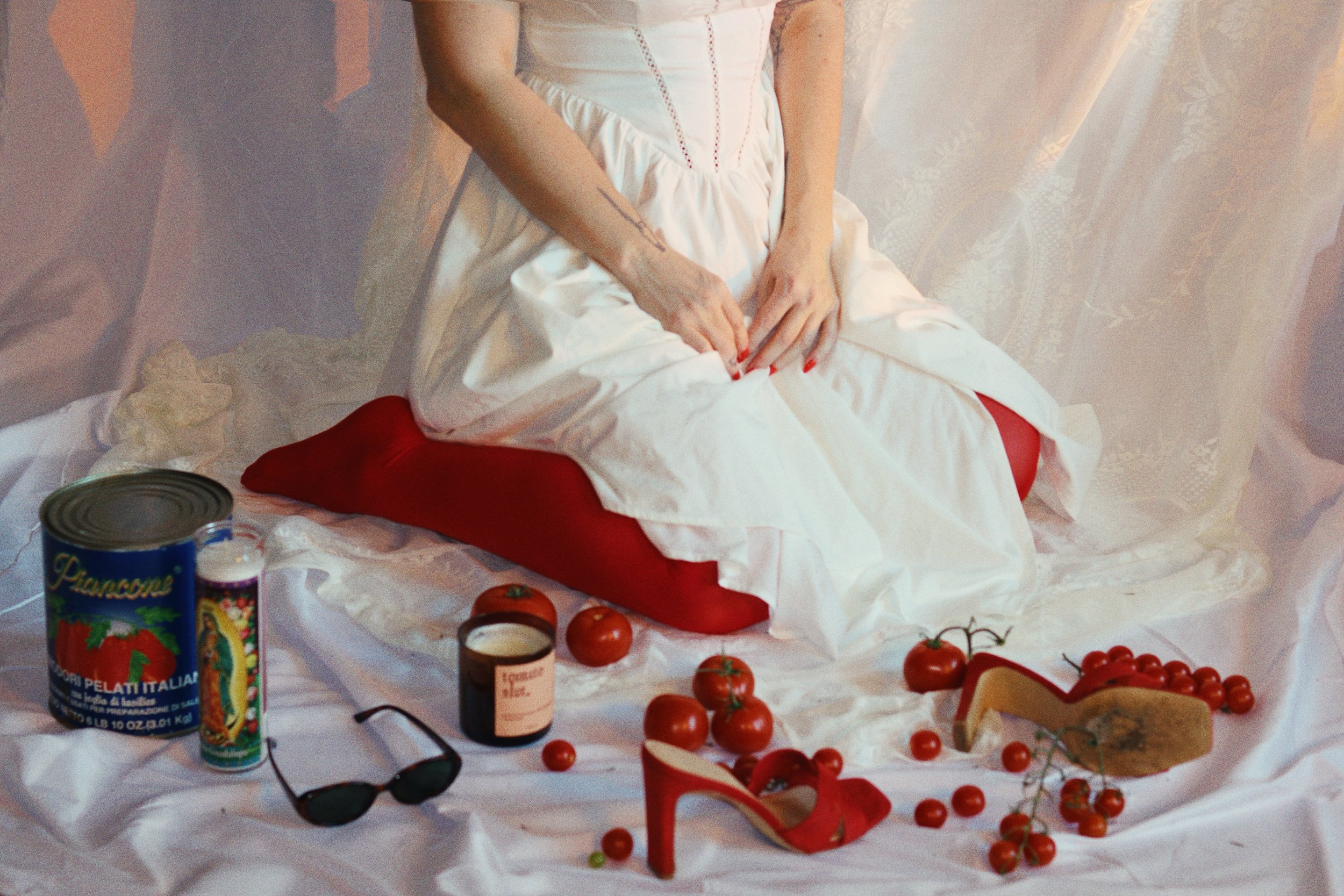 A woman with red-painted nails sits on a red cushion, wearing a white dress with red stitching, surrounded by tomatoes, a pair of sunglasses, a can of Italian spaghetti, a small jar, a wedge of cheese, and red high-heeled shoes on a white cloth backg
