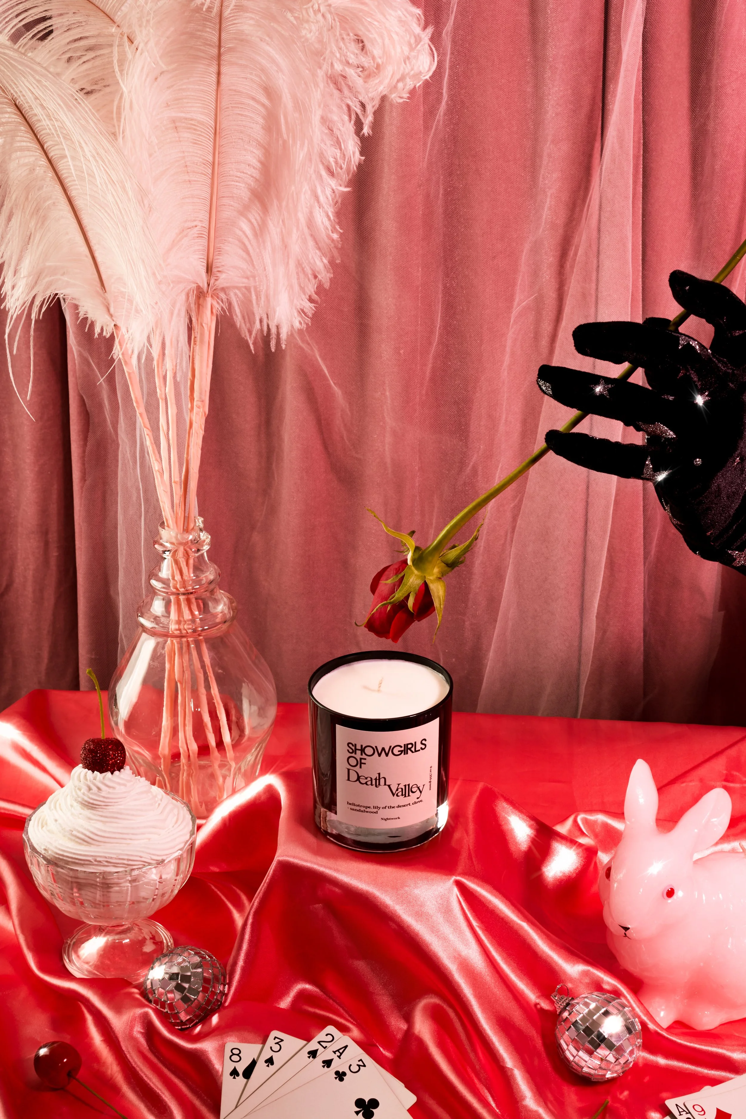 A Valentine's Day themed still life scene with pink feathers in a glass vase, a red rose, a lit scented candle labeled "SHOWGIRLS OF Death Valley," a white ceramic rabbit, a bowl of whipped cream with a cherry on top, playing cards, and small disco b