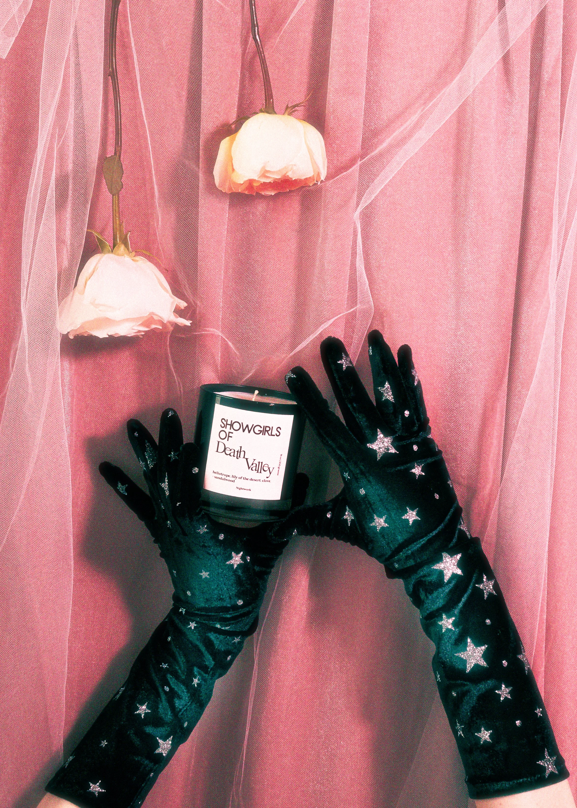 Person wearing star-patterned black gloves holds a black candle labeled "Showgirls of Death Valley" against a pink background with hanging pink roses and sheer fabric.