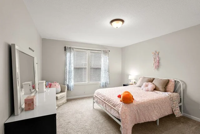 A bedroom with a bed, plush toys, a window with curtains, a bedside lamp, a white dresser, a mirror, and wall decor.