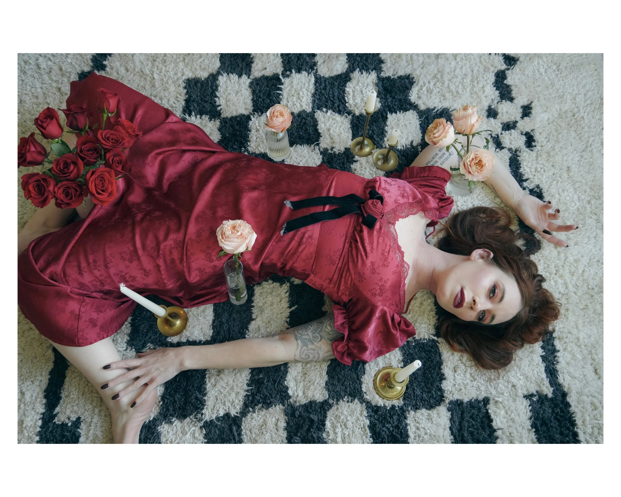 A woman with red hair lying on a black and white patterned rug surrounded by flowers and candlesticks.