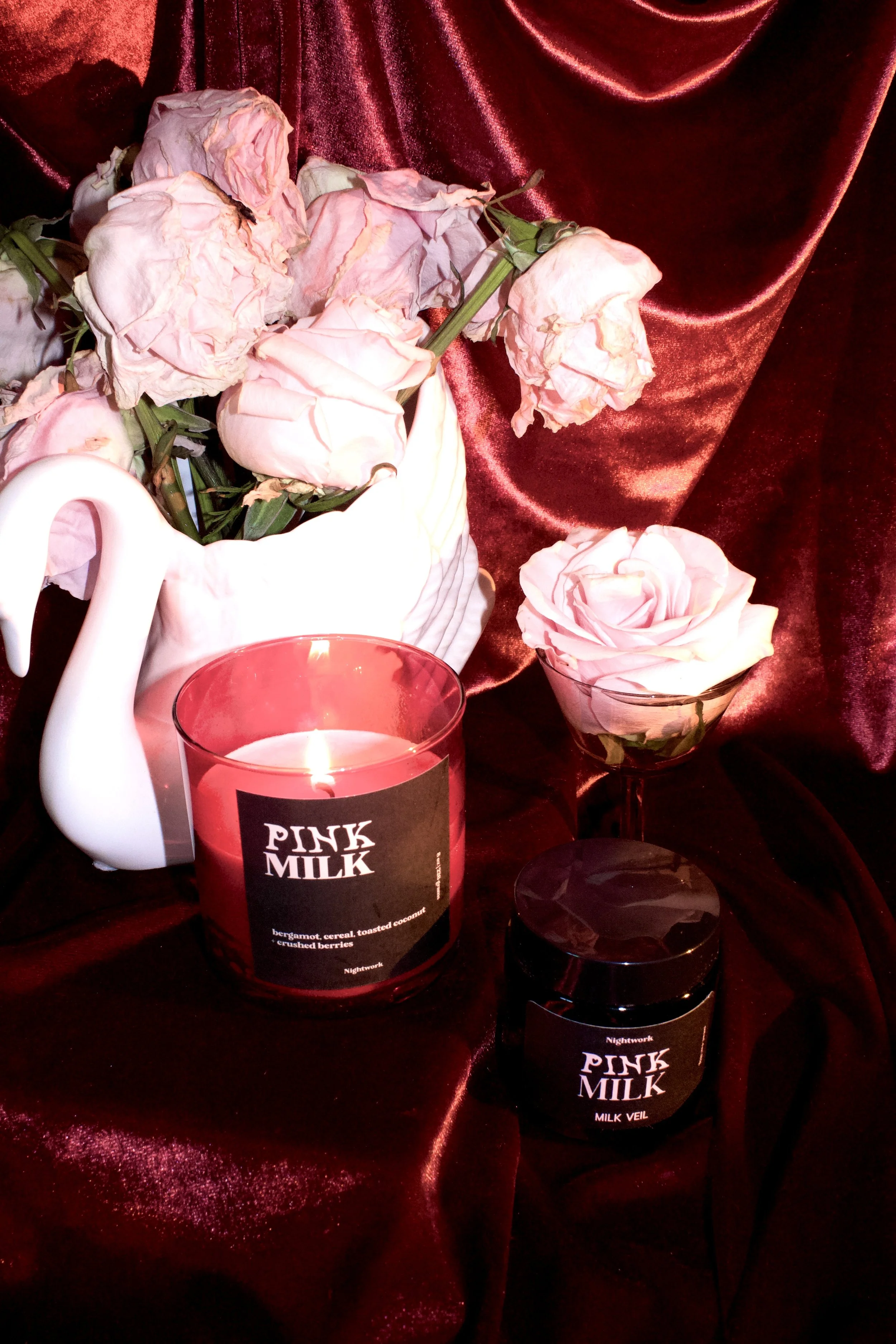 A still life arrangement featuring a bouquet of wilted pink roses in a white ceramic vase, a single fresh pink rose in a glass bowl, a lit pink candle labeled 'Pink Milk,' and a small black container labeled 'Pink Milk Milk Veil,' all set against a d