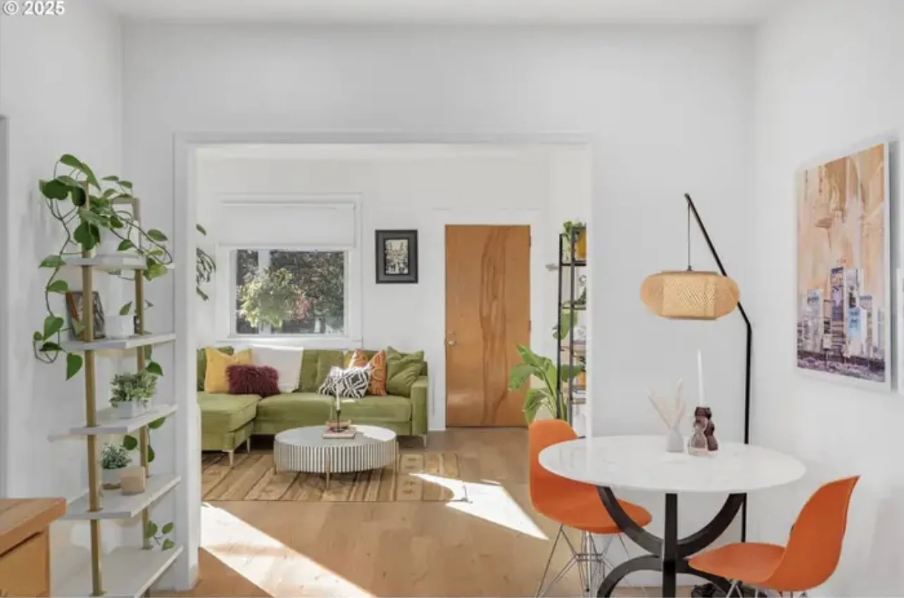 Bright living room with a white wall, green sectional sofa with colorful pillows, and a wooden door. Adjacent is a small dining area with an oval white table, orange chairs, and a black and wood standing lamp. There are decorative shelving units with