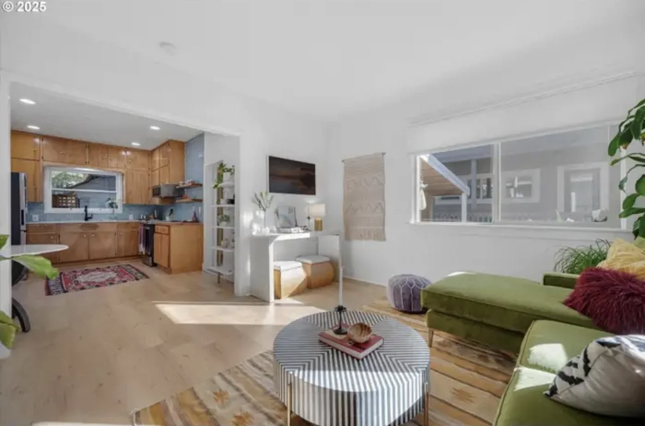 Bright living room with green sofa, colorful pillows, coffee table, and large windows; open kitchen with wooden cabinets and a window over the sink.
