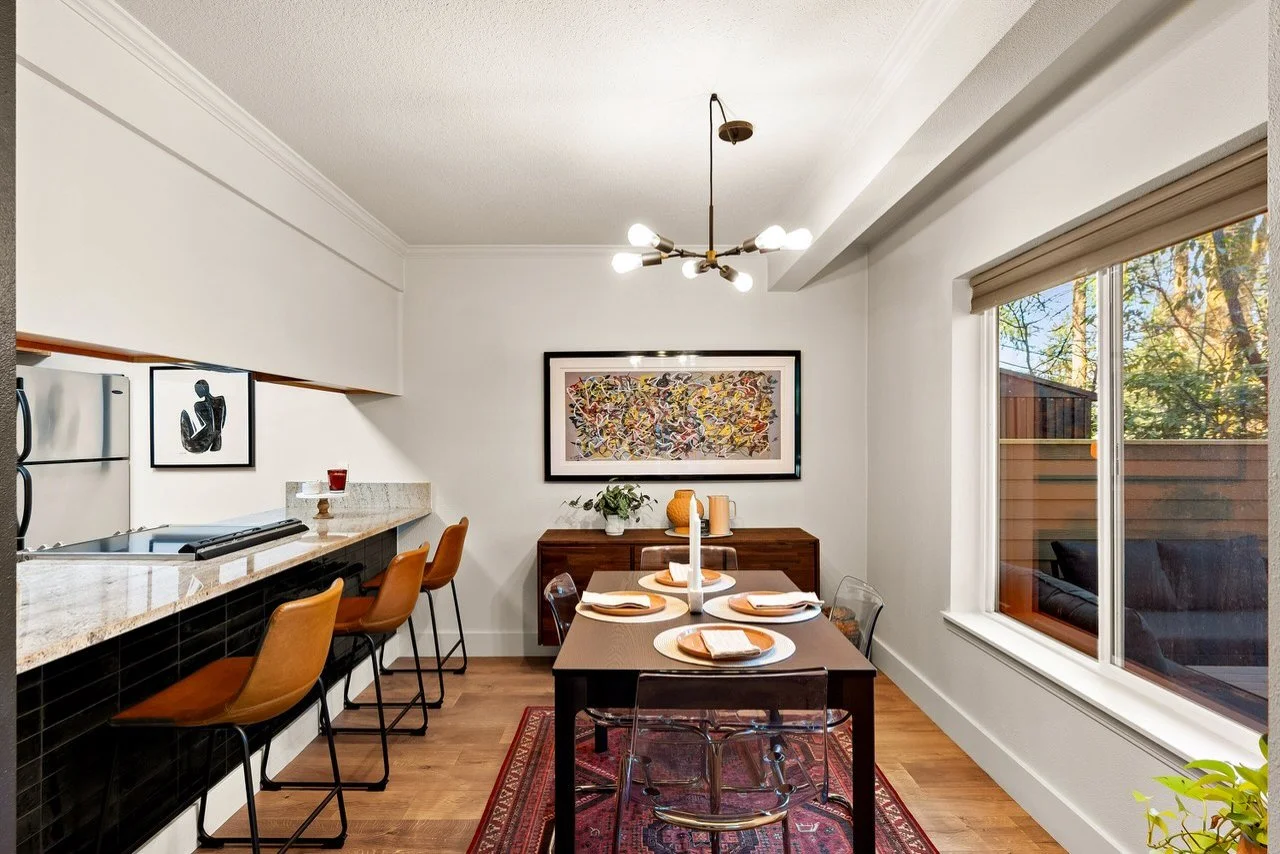 Dining room with a dark wood table set with white plates, napkins, and a candle centerpiece, a sideboard with plants and decorative items, and a large window showing trees outside, with modern light fixture on the ceiling.