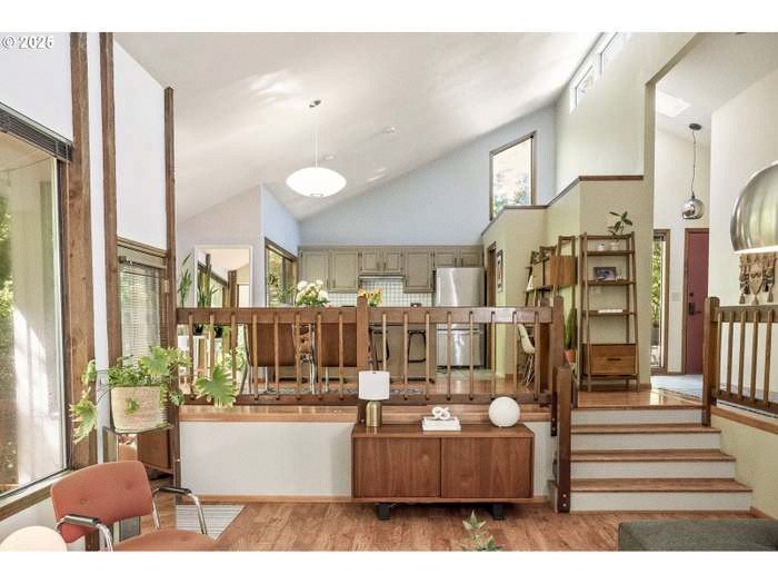 Living area with wooden railings and steps leading to kitchen, large windows with sunlight, plants, and modern decor.