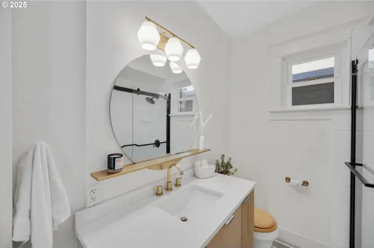 Modern bathroom with a white vanity, gold fixtures, a round mirror, a small potted plant, a white towel hanging, and a window above the toilet, with a black sliding barn door shower visible in the mirror reflection.