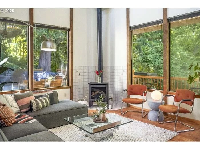 Living room with large windows, a gray sectional sofa with pillows, two orange chairs, a glass coffee table with plants, a white rug, and a small black fireplace, with green trees visible outside.