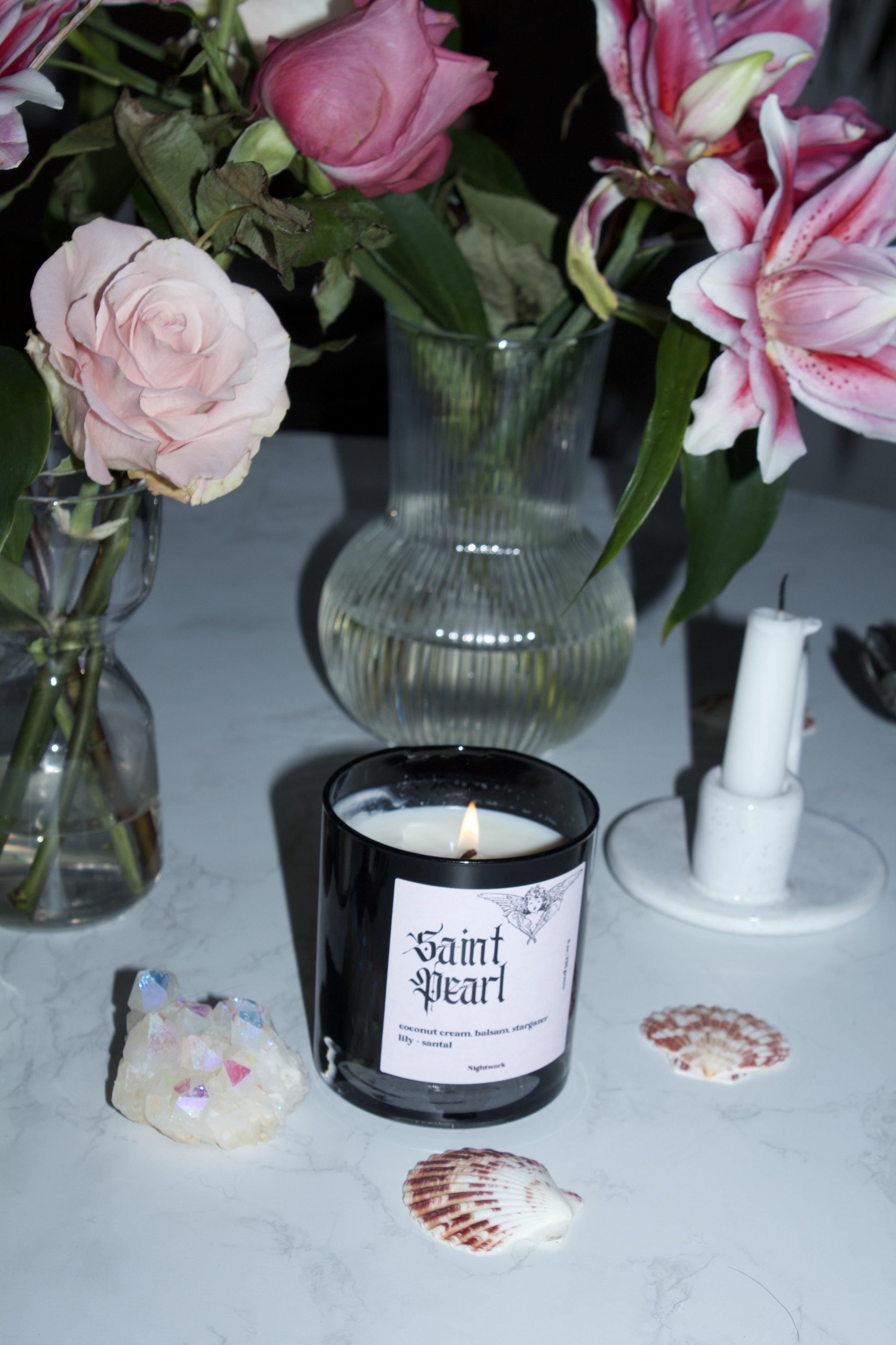 Vase with pink roses and pink lilies, lit Saint Pearl candle on white marble surface with seashells and a candle holder.