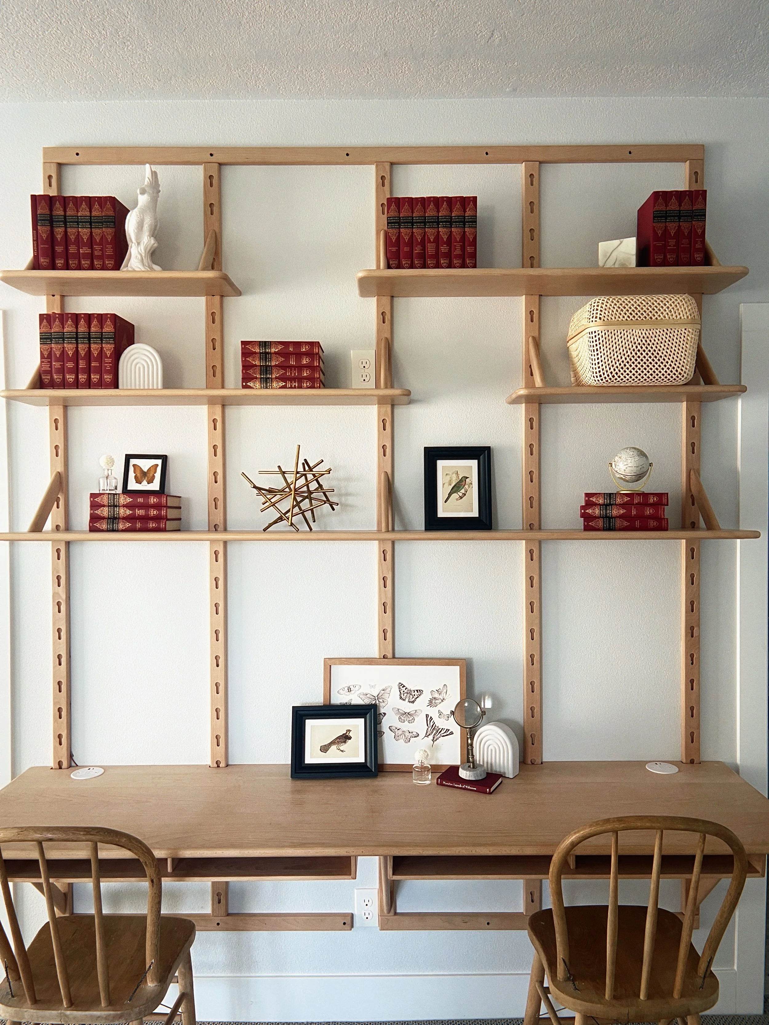 Interior wall with wooden bookshelves holding red books, decorations, framed pictures, and a desk with two wooden chairs below.