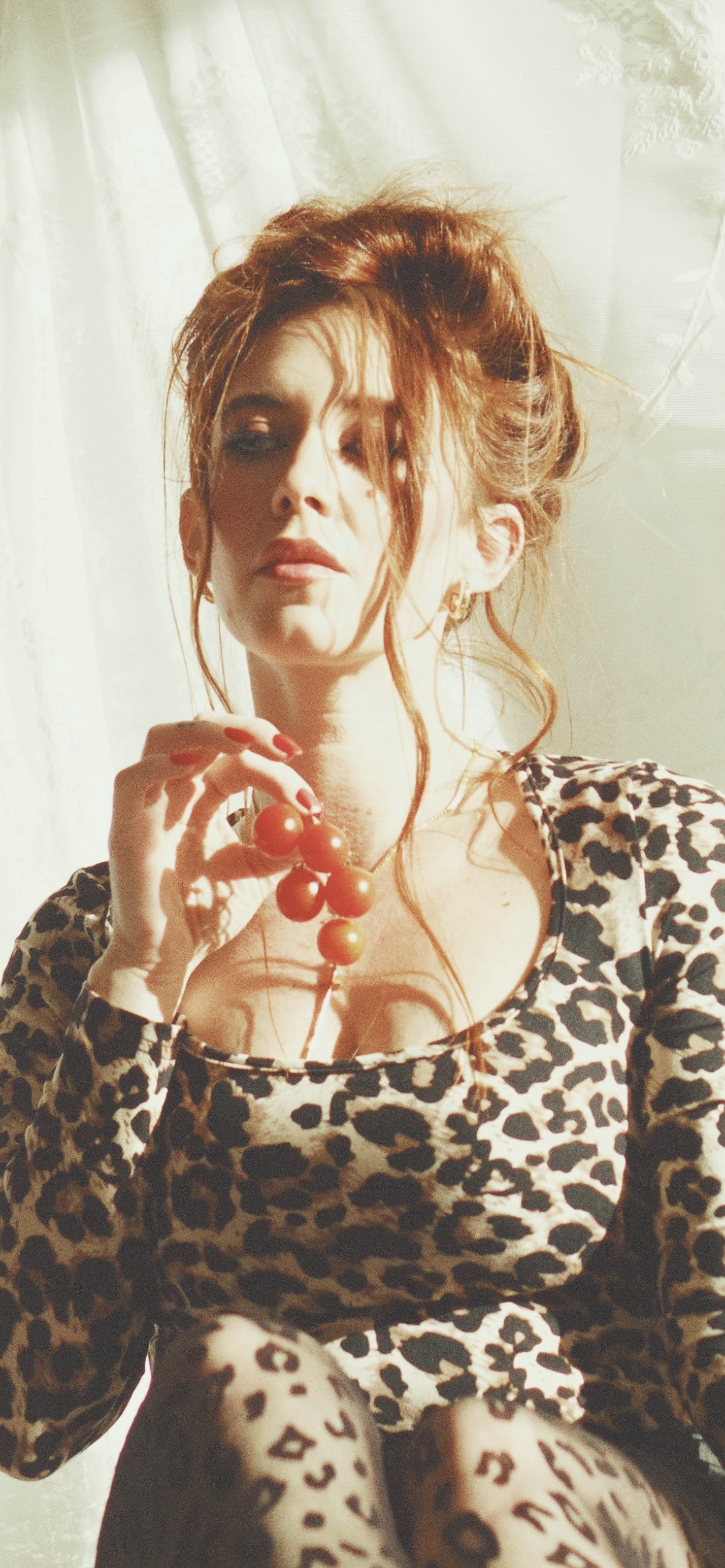 A woman with red hair styled in an updo, wearing a leopard print top and leggings, holding a string of orange beads, and sitting near a window with sheer white curtains.