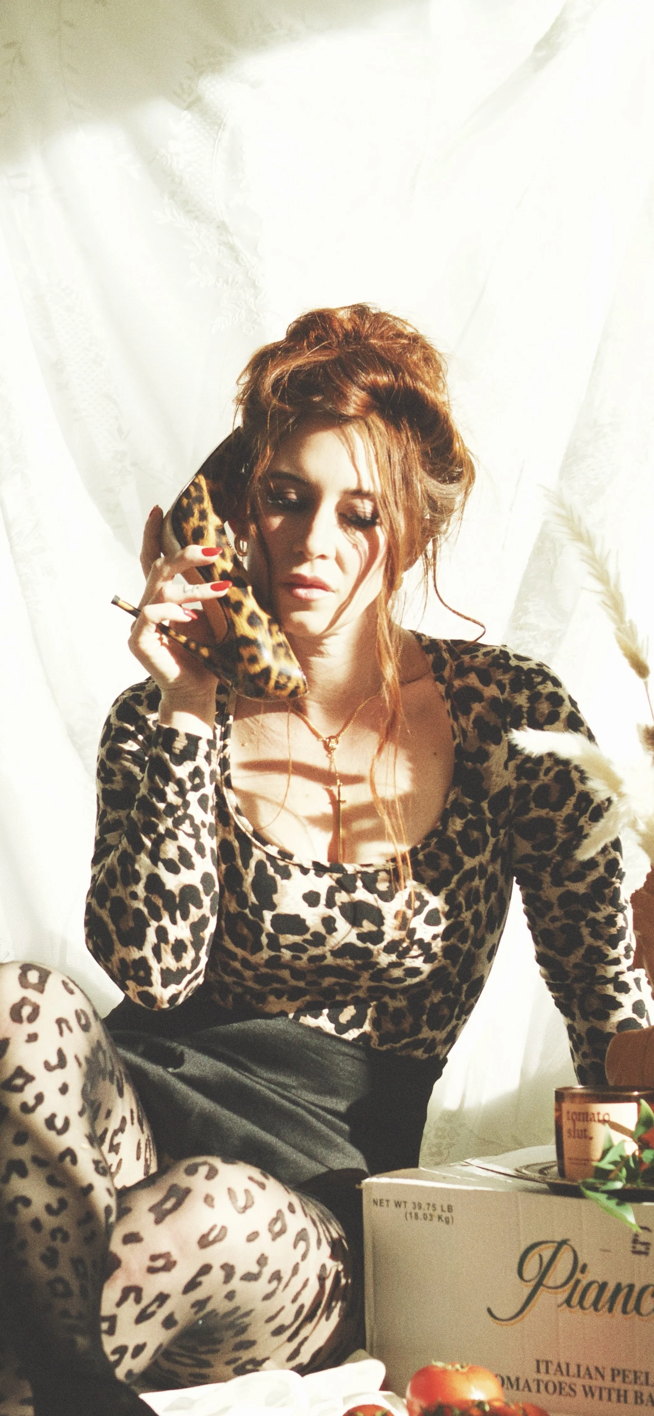 A woman with red hair wearing a leopard print top and leopard print legging, sitting indoors, talking on a leopard print telephone, with a white curtain background and some food items on the table in front of her.