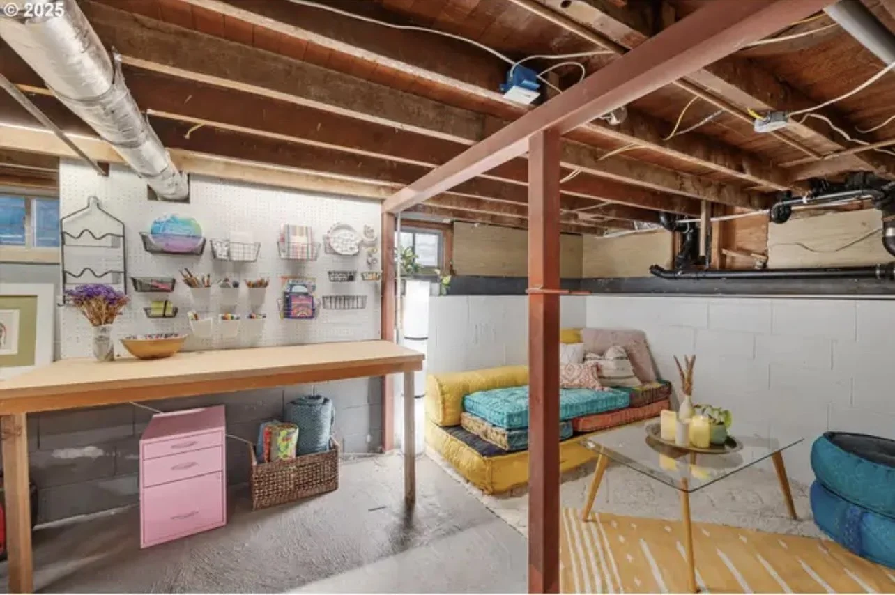 A basement room with unfinished wooden ceiling beams, a pegboard wall with storage baskets, a work table, colorful cushions on a low seating area, and a small glass table with candles and decor, featuring exposed pipes and a window.