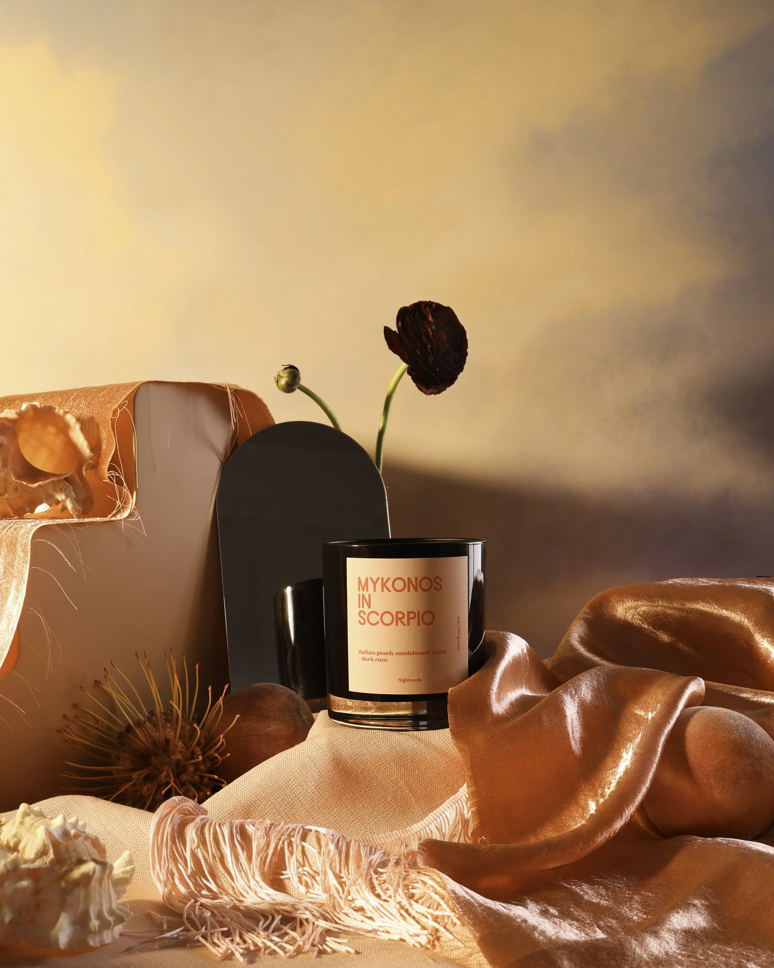 A black candle labeled 'Mykonos in Scorpio' with a pink background, surrounded by silk fabrics, flowers, and decorative objects, against a soft sunset sky.
