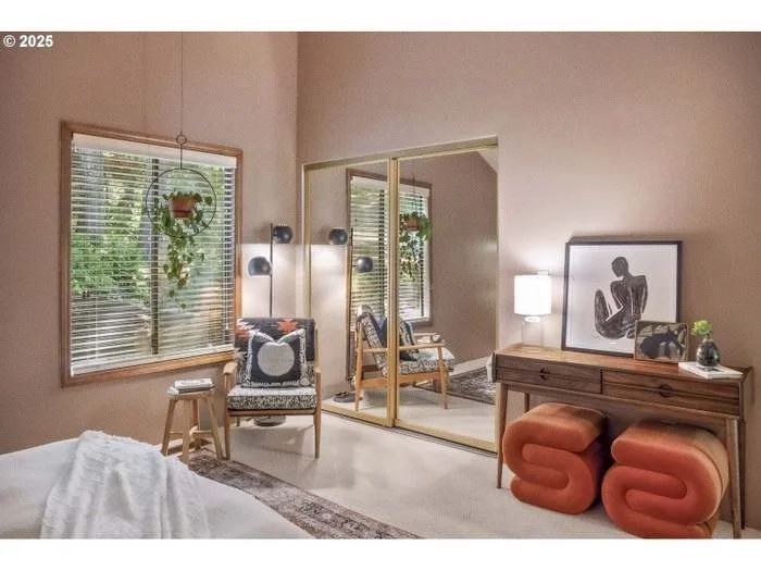 A cozy bedroom with a large window, sitting area, and mirrored closet. Decor includes artwork, a lamp, and two orange ottomans.