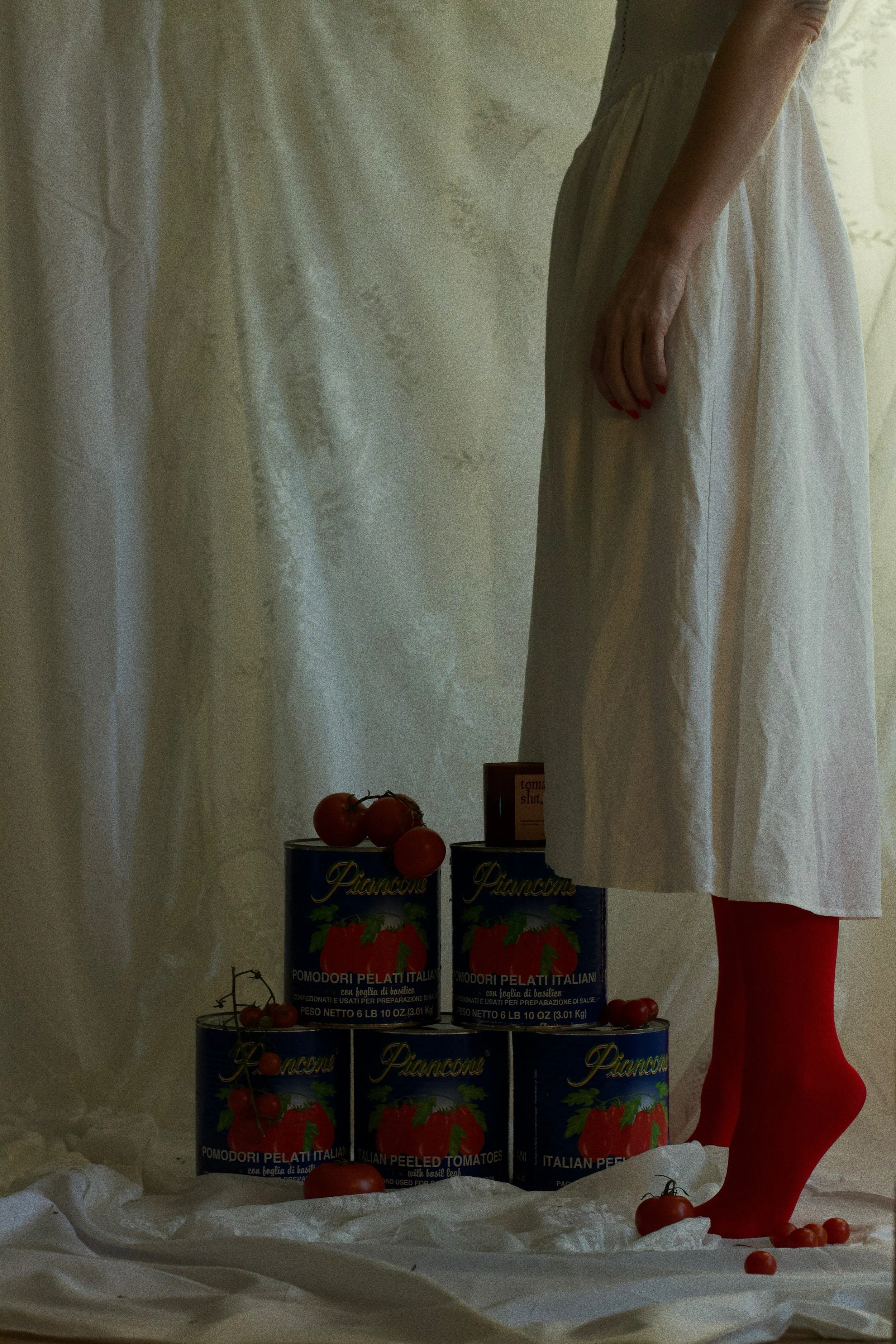 A person wearing a white dress and red tights stands beside cans of peeled Italian tomatoes, with other tomatoes scattered on the fabric beneath them. Only the lower half of the person's body is visible.