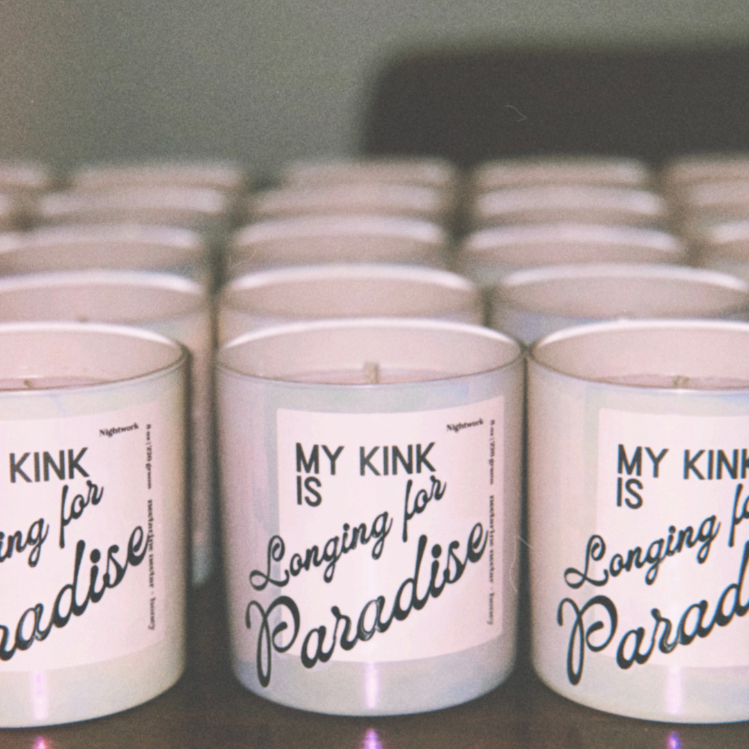 Grouped candles with white labels that read 'KINK is Longing for Paradise' on a shelf.