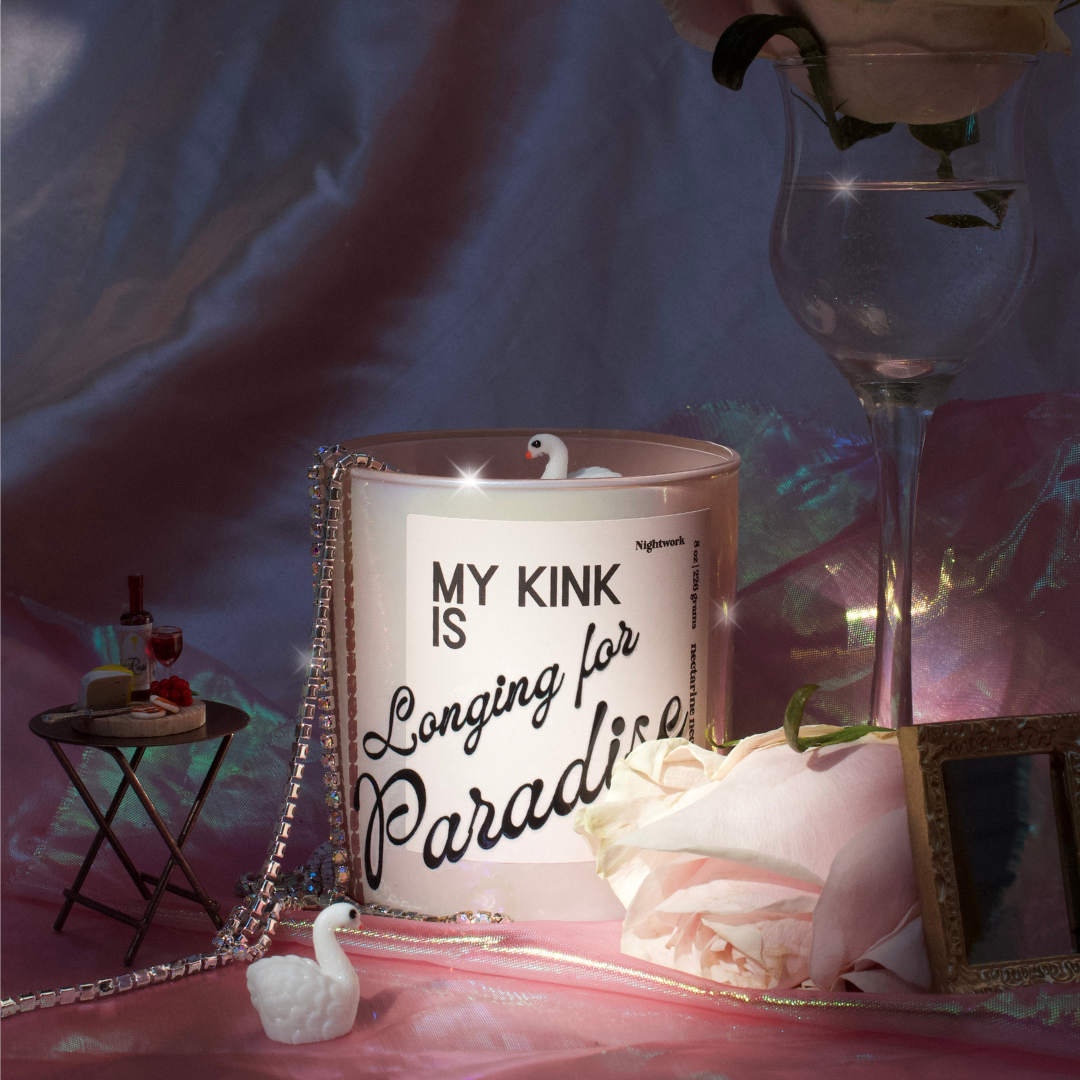 A decorative scene with a mug that has the phrase 'My Kink is Longing for Paradise' on it, a small swan figurine, a pink rose, a tiny picture frame, and various small items arranged on a pastel-colored surface with a multicolored, iridescent backdrop