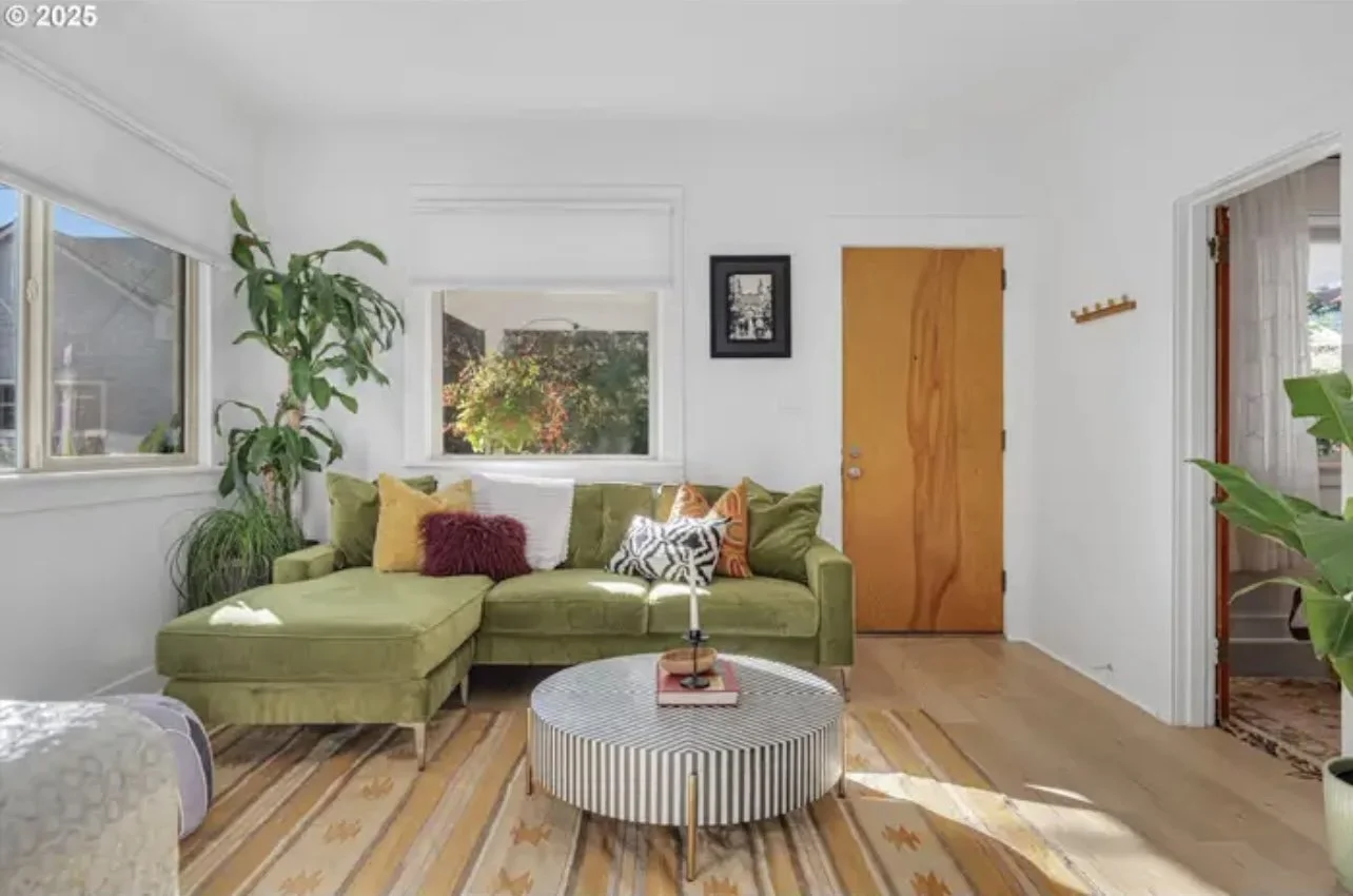 Living room with a green sectional sofa, colorful pillows, large window, wooden door, round striped coffee table, potted plants, and framed black-and-white photo on white walls.