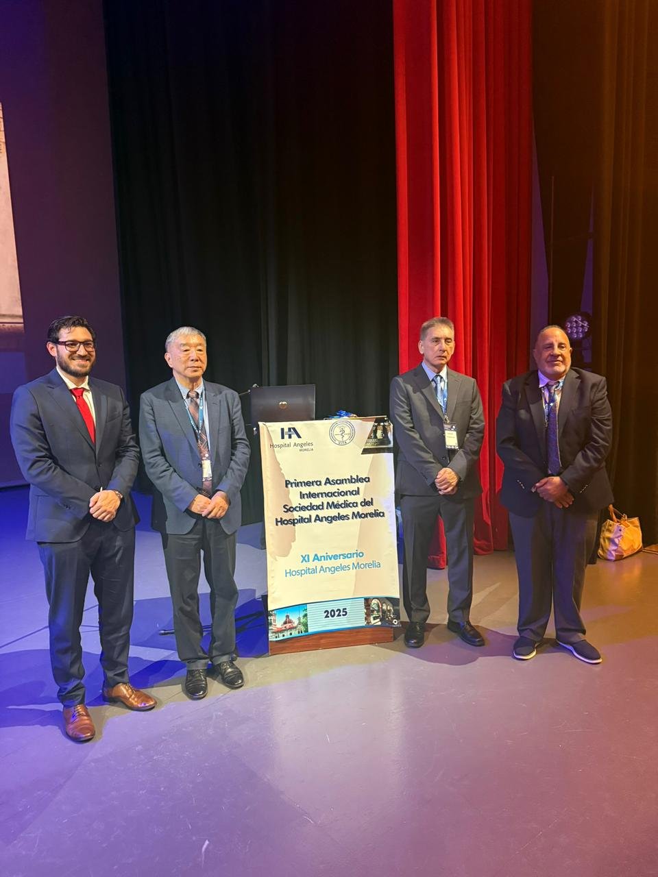 Cuatro hombres de negocios en traje de pie junto a un podio con cartel que dice 'Primer Asamblea Internacional Sociedad Médica del Hospital Angeles Morelia' en un auditorio con cortinas rojas y negras.