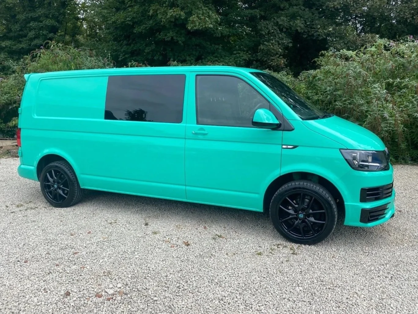 A teal-colored van with black wheels parked on gravel, with trees and bushes in the background.